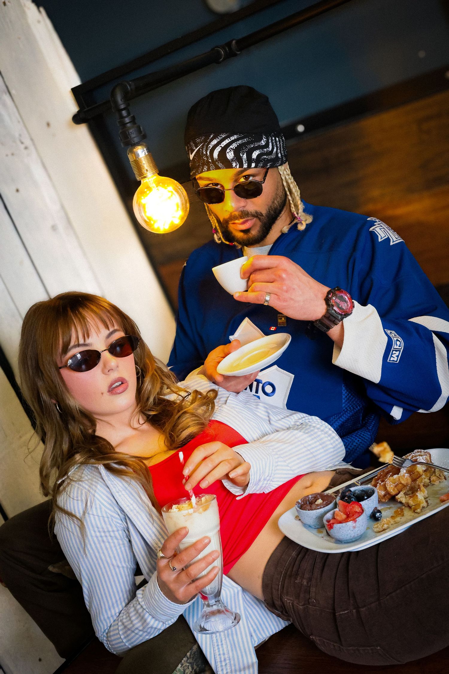 Two stylish people wearing sunglasses enjoy drinks and food together in a trendy restaurant setting.