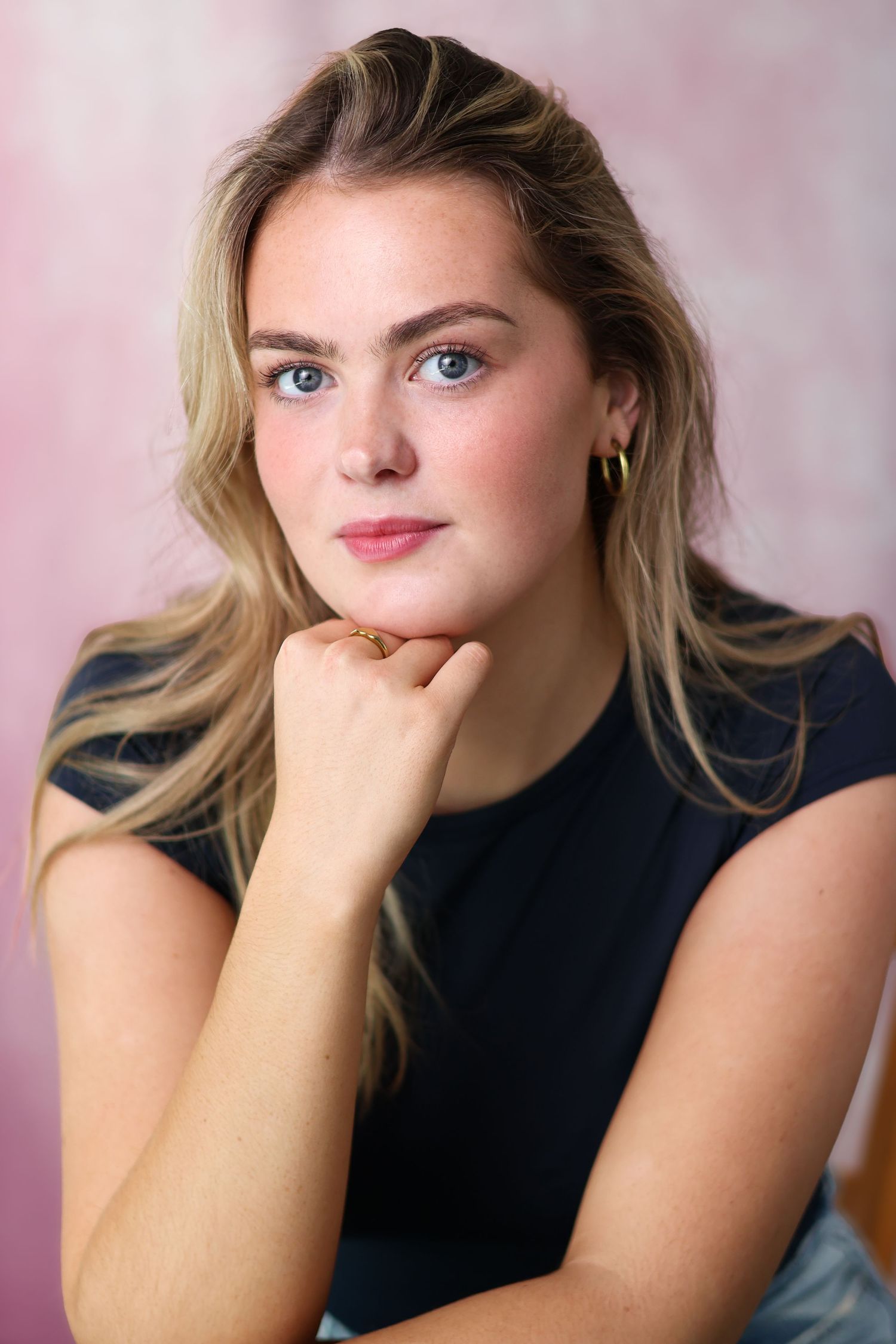A person with blonde hair poses against a pink backdrop in a navy top while resting their chin on their hand.