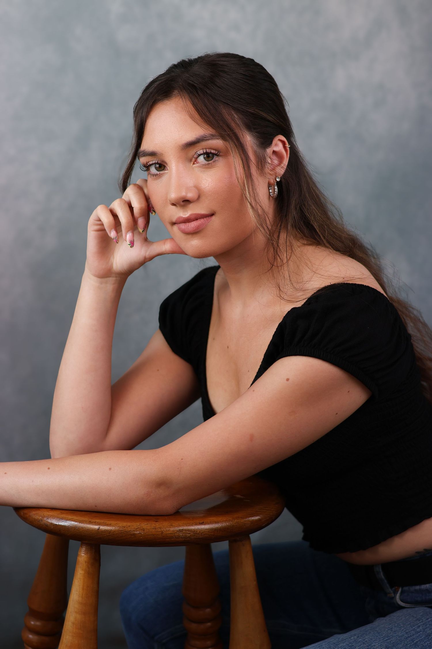 Young woman in black off-shoulder top poses casually while sitting on wooden stool against gray backdrop.