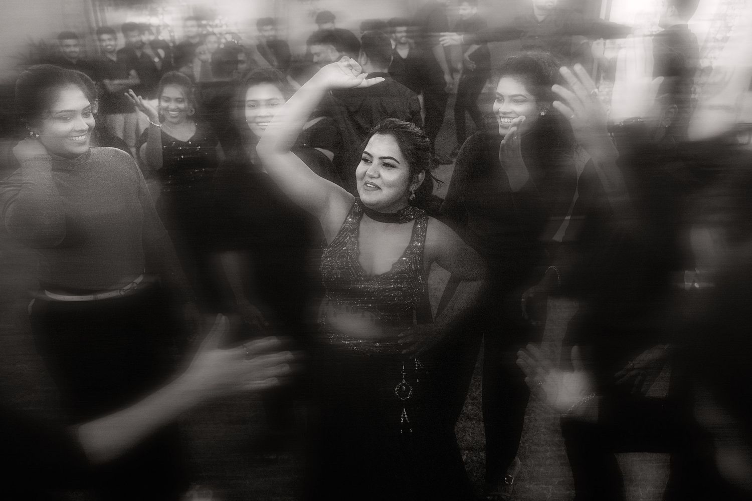 Black and white photo of a woman dancing joyfully in a crowded party, arms raised, surrounded by blurred figures.
