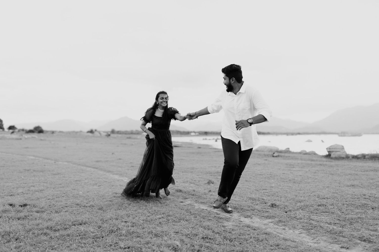 Black and white photo of a couple dancing joyfully outdoors near a lake, woman in flowing dress, man in white shirt.