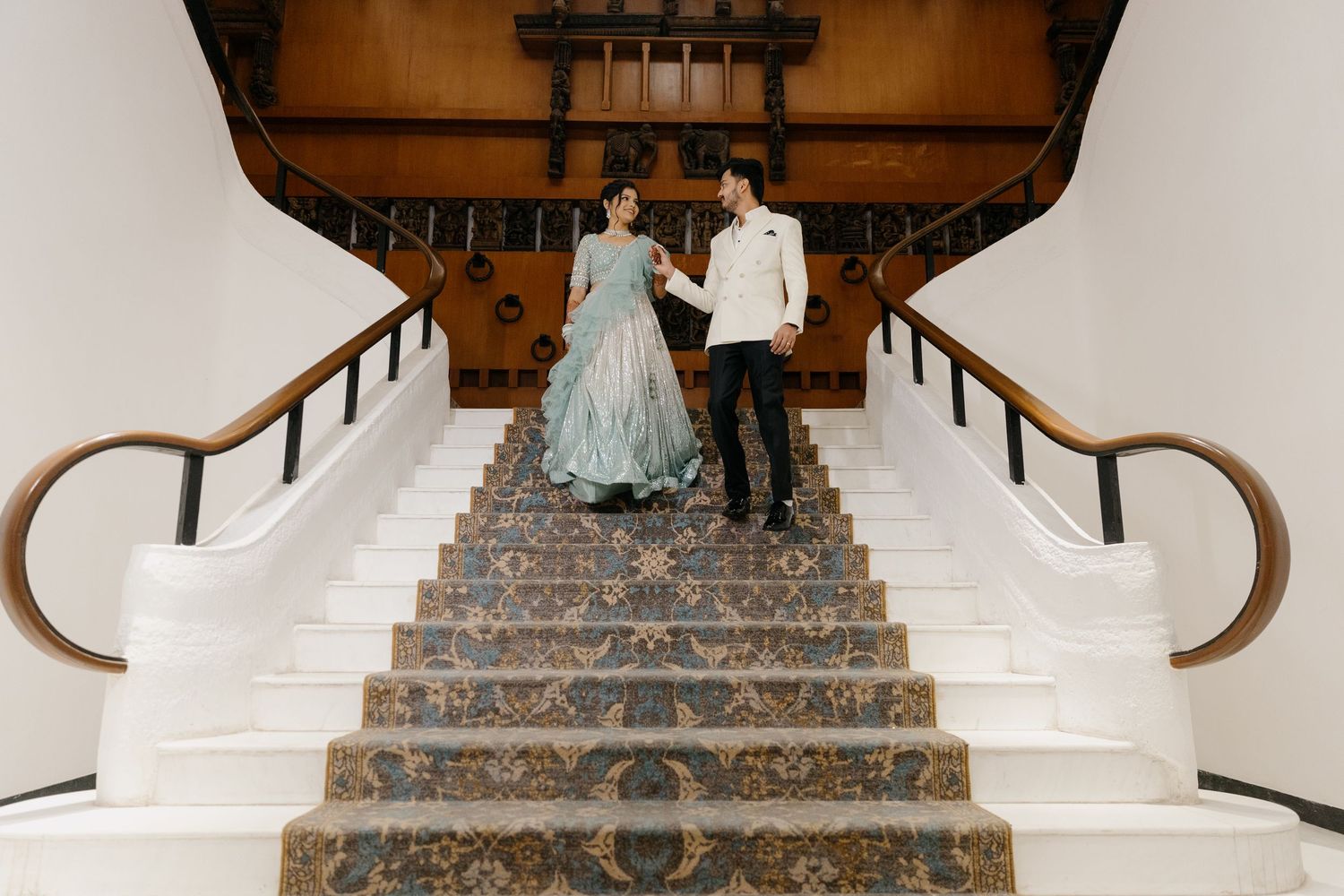 Couple in formal attire descending an elegant staircase with ornate carpet and wooden railings.