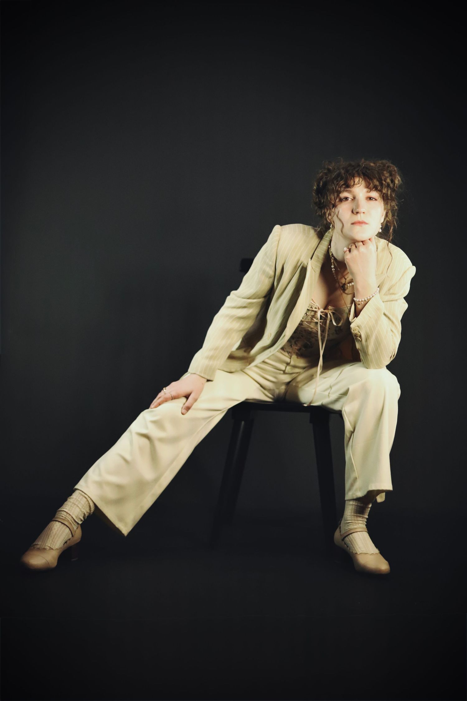 A fashion portrait of someone in a beige suit sitting on a chair against a dark background.