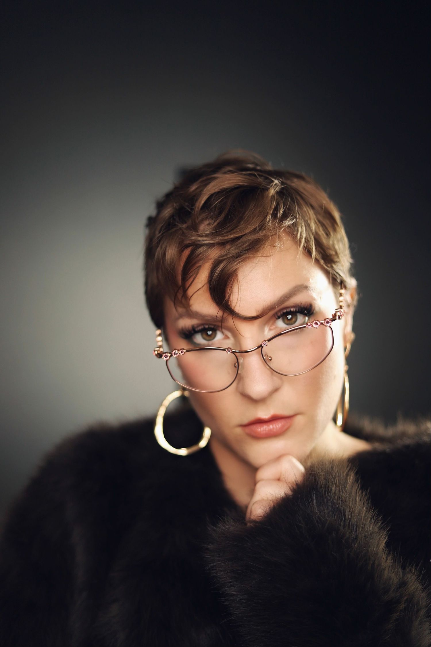 Stylish portrait featuring round glasses, gold hoop earrings, and fur coat against dark background.