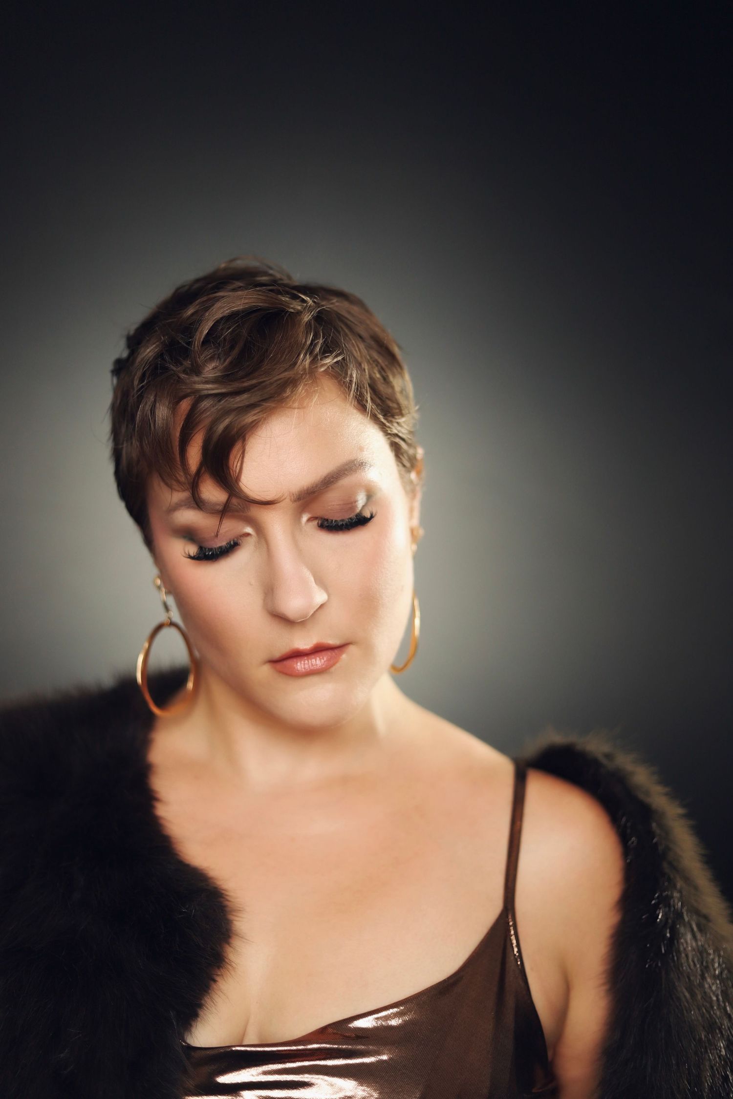 Glamour portrait of a person in fur coat with short hair and gold hoop earrings against gradient background.