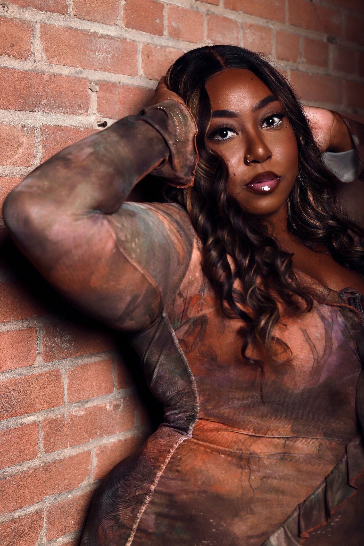 A woman with long curly hair leans against a brick wall wearing an artistic earth-toned bodysuit with tattoo-like designs.