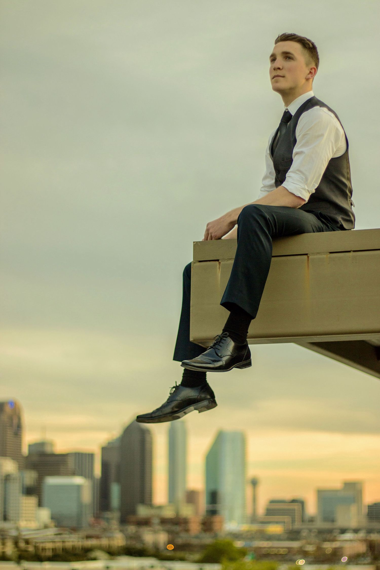 Person in business attire sitting on ledge overlooking city skyline at sunset.