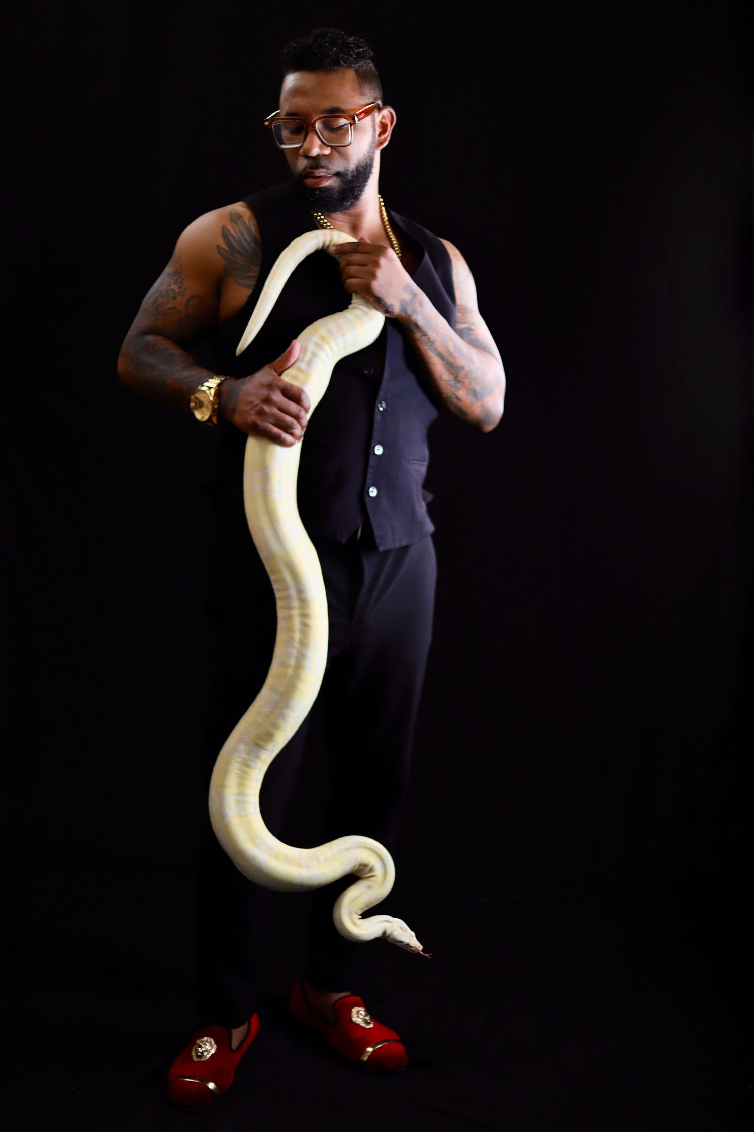 Man in black vest holding a large albino python snake, wearing red loafers and gold watch against dark background.