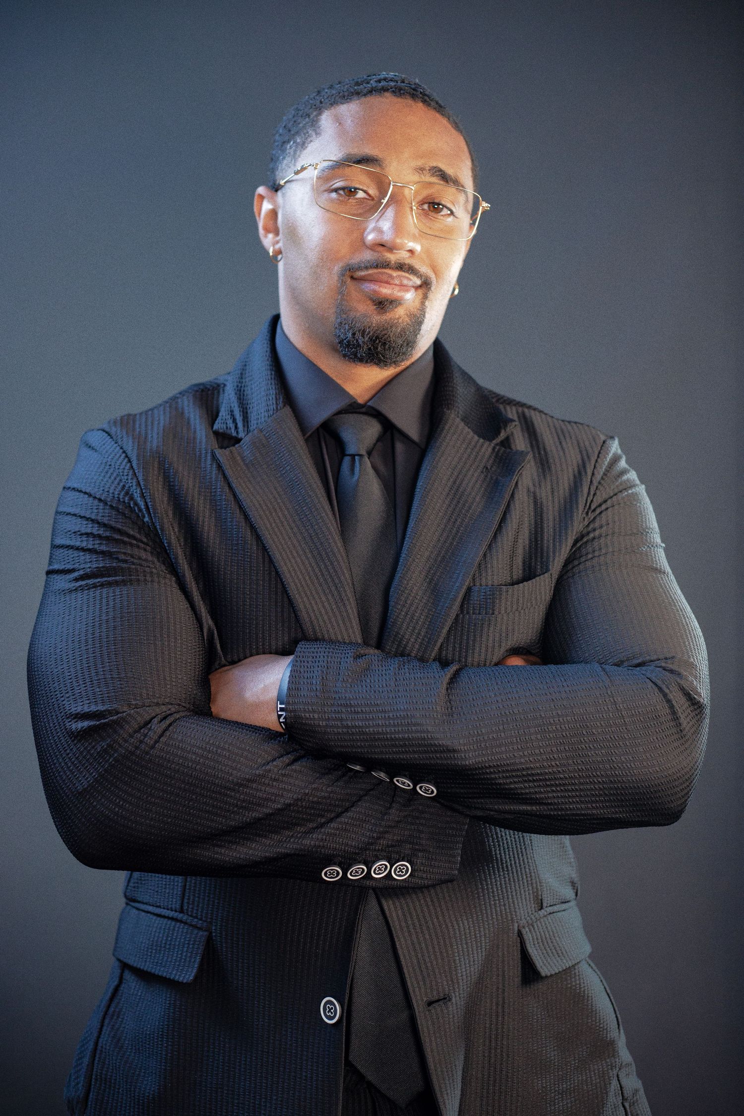 Professional Black man in pinstripe suit with arms crossed, wearing glasses and earring, against gray background.