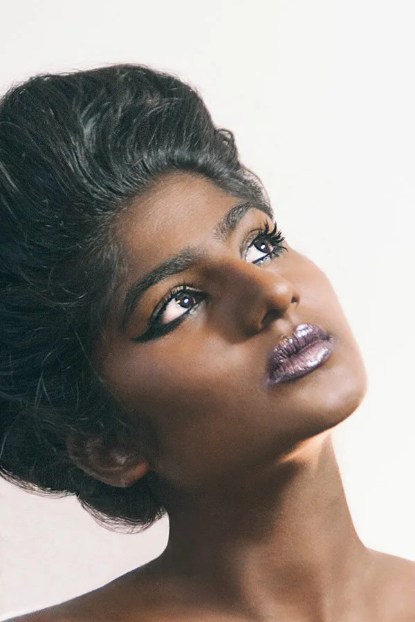 Portrait showing dramatic makeup with purple lipstick and long lashes against white background.