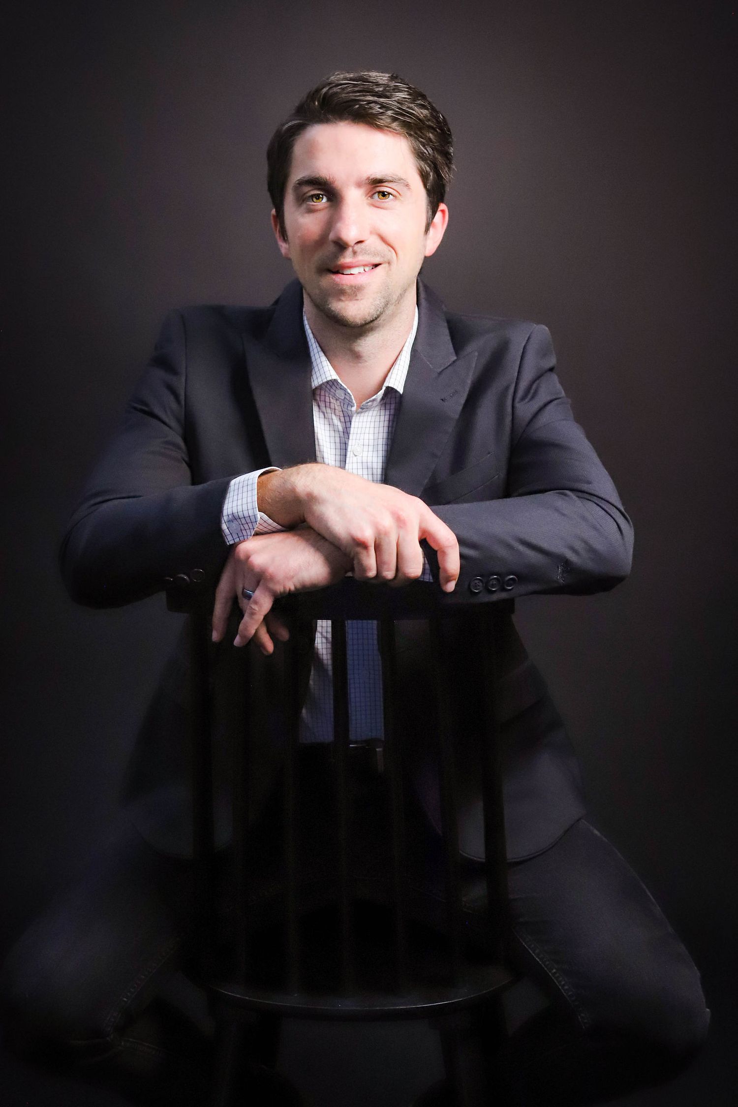 Professional studio portrait of someone in a dark suit sitting backwards on a chair against a black background.