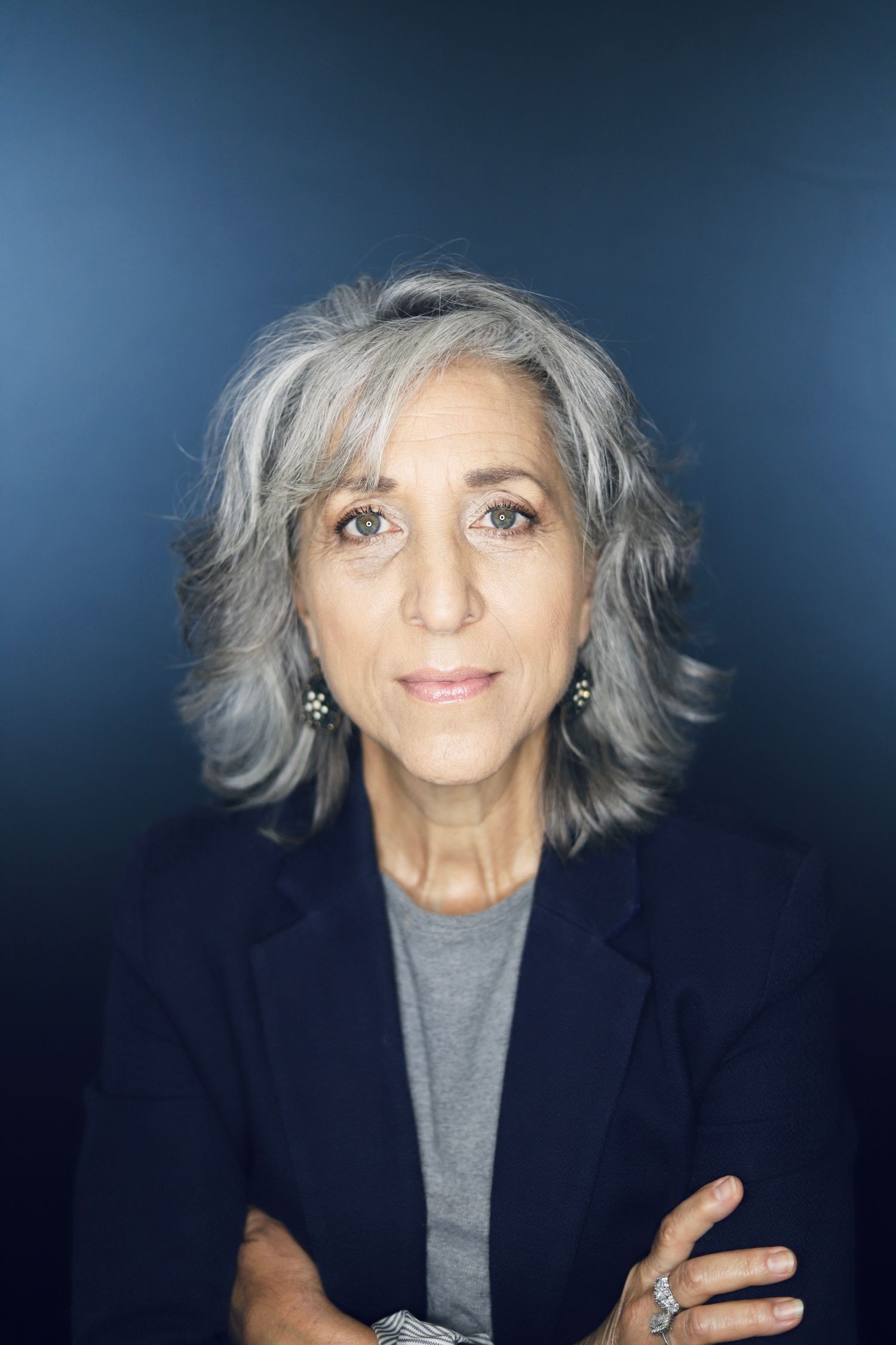 Professional headshot against blue background wearing navy blazer and grey top.
