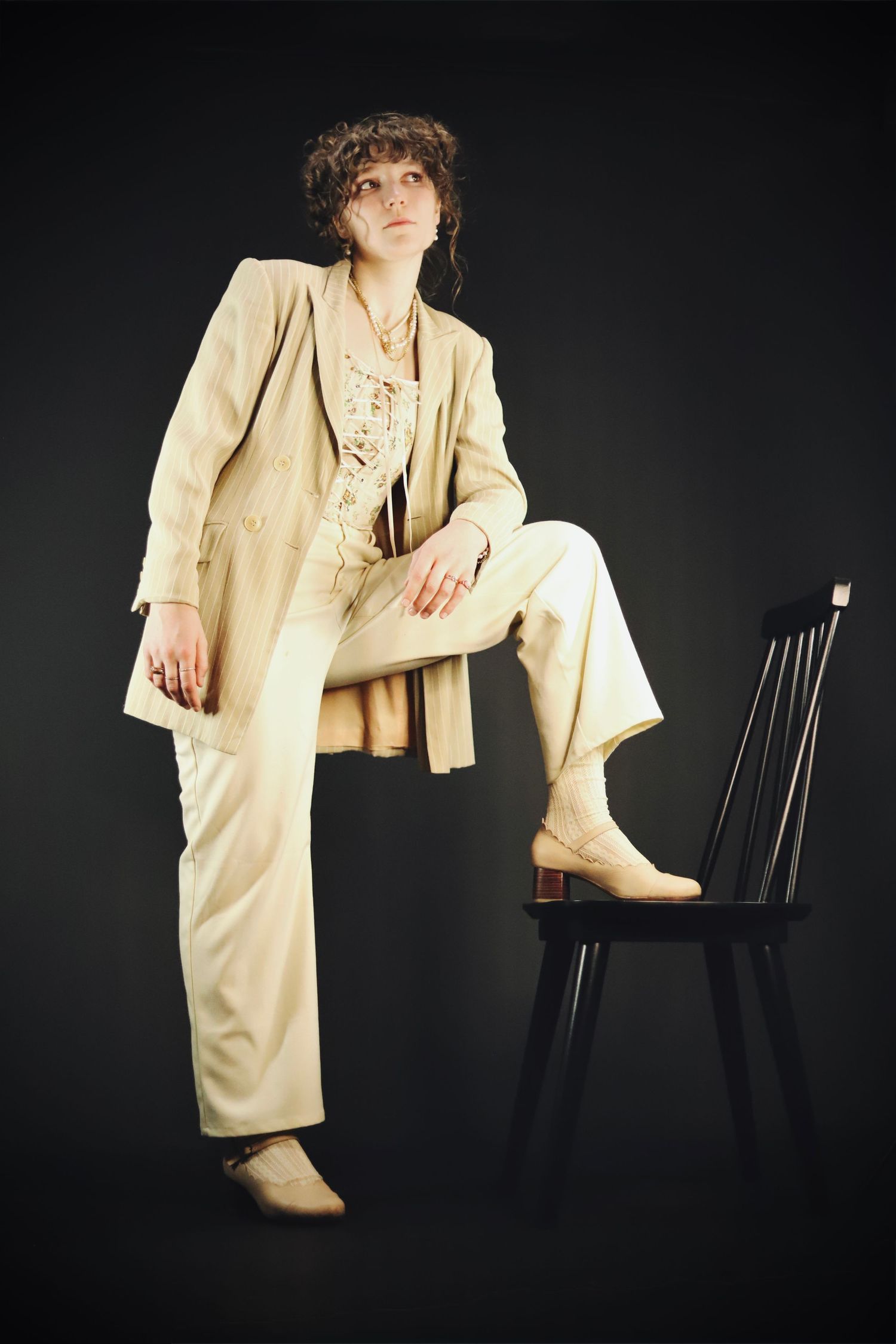 Artistic fashion portraits of a person in beige suit posing dramatically with a black chair against dark backdrop.