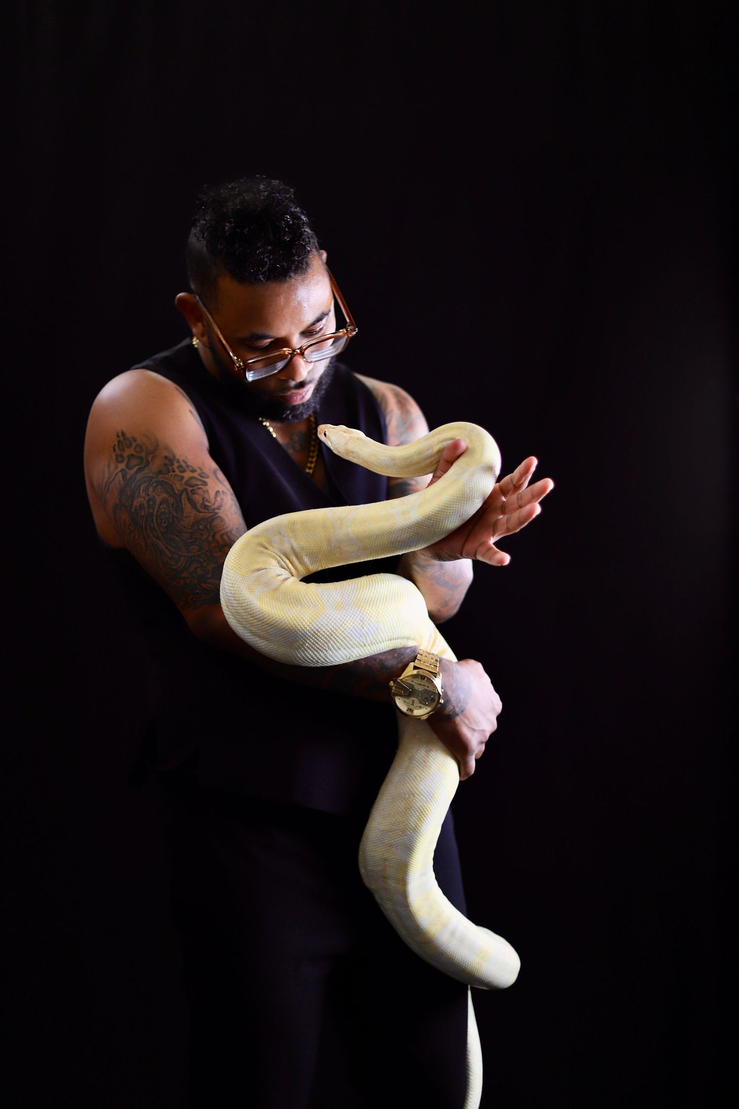 Tattooed man in black sleeveless shirt holding a large albino python snake against dark background.