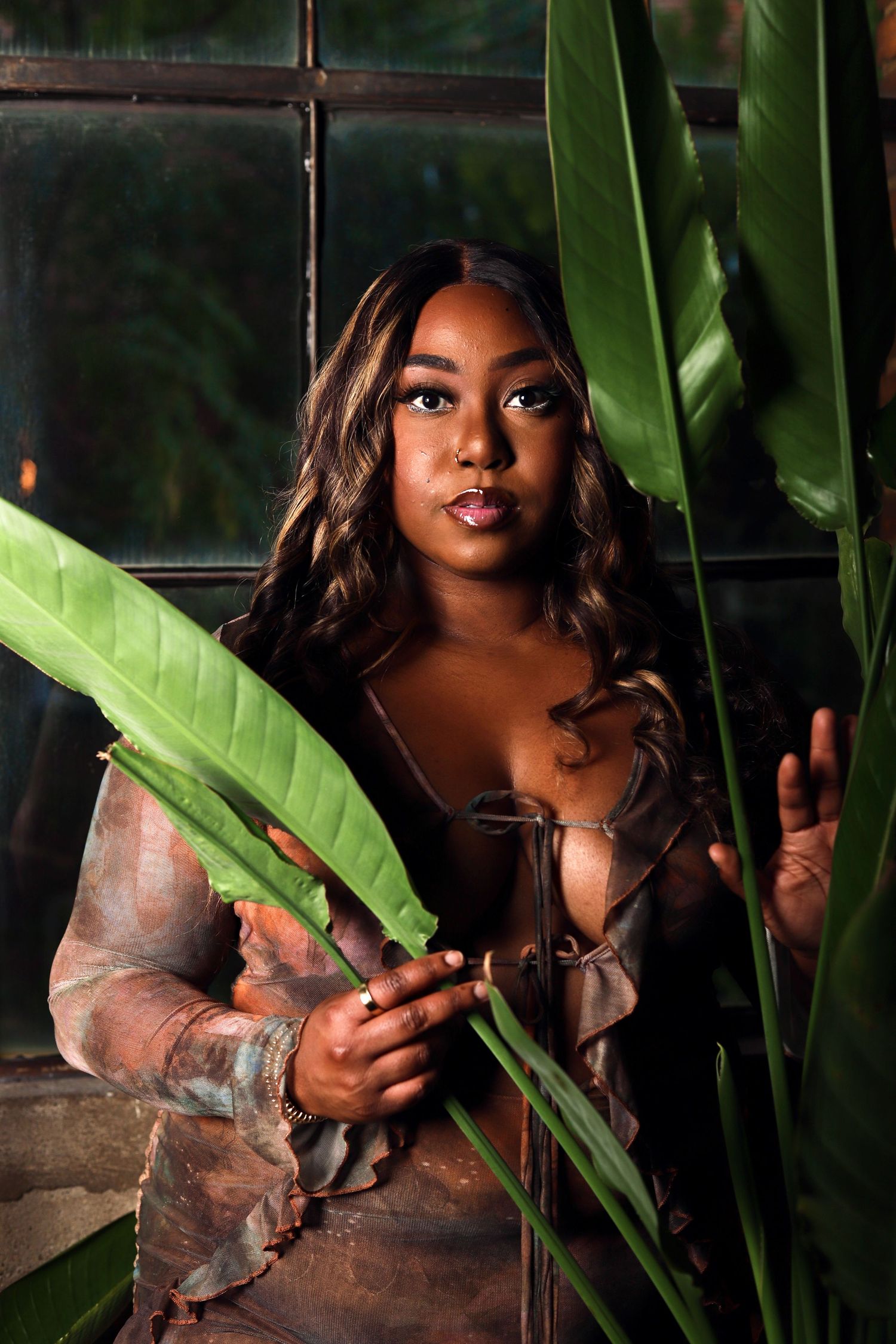 A young Black woman poses among tropical green leaves wearing a sheer brown top, with dark windows in the background.