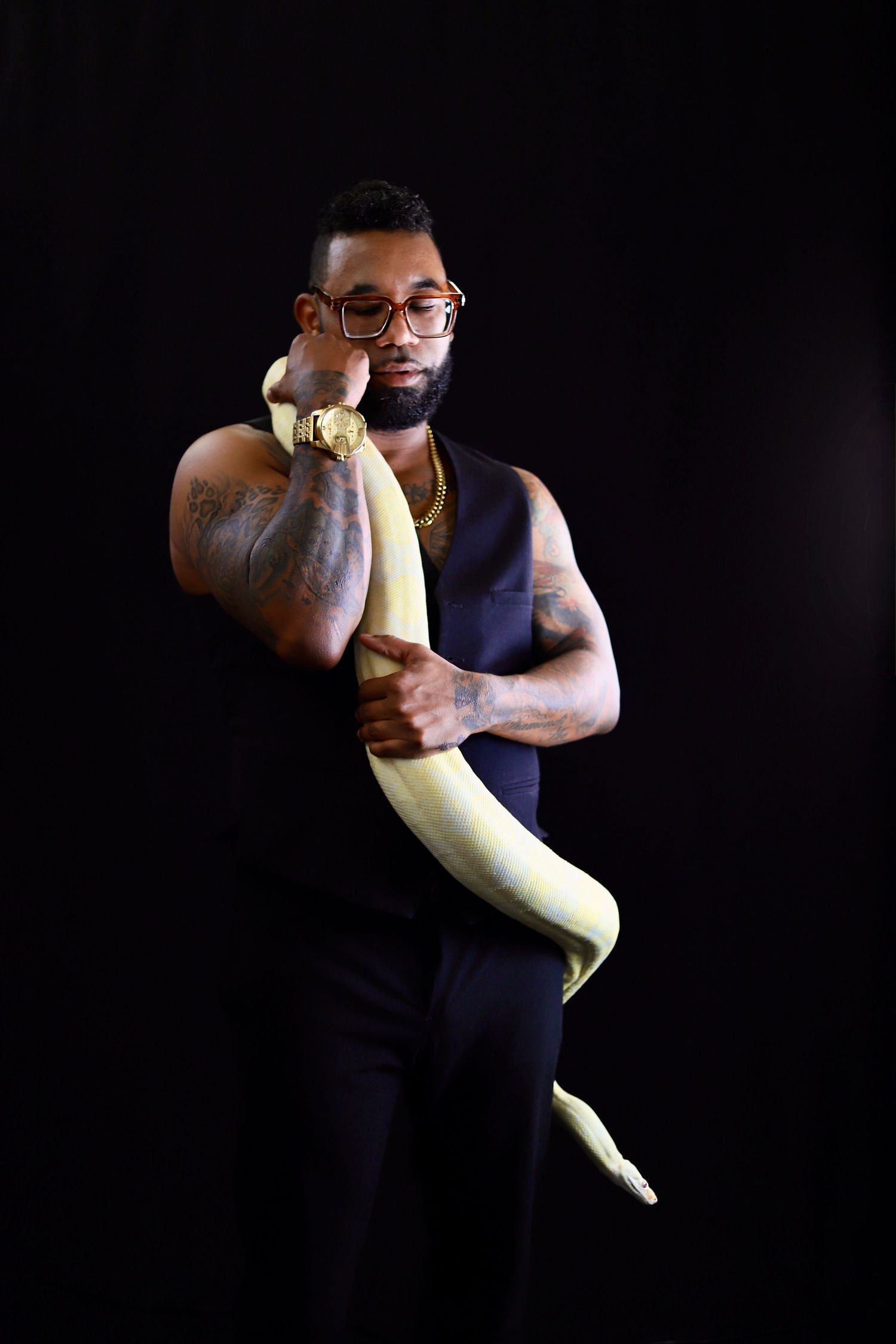 Tattooed man with glasses holding a yellow albino snake against a dark background.