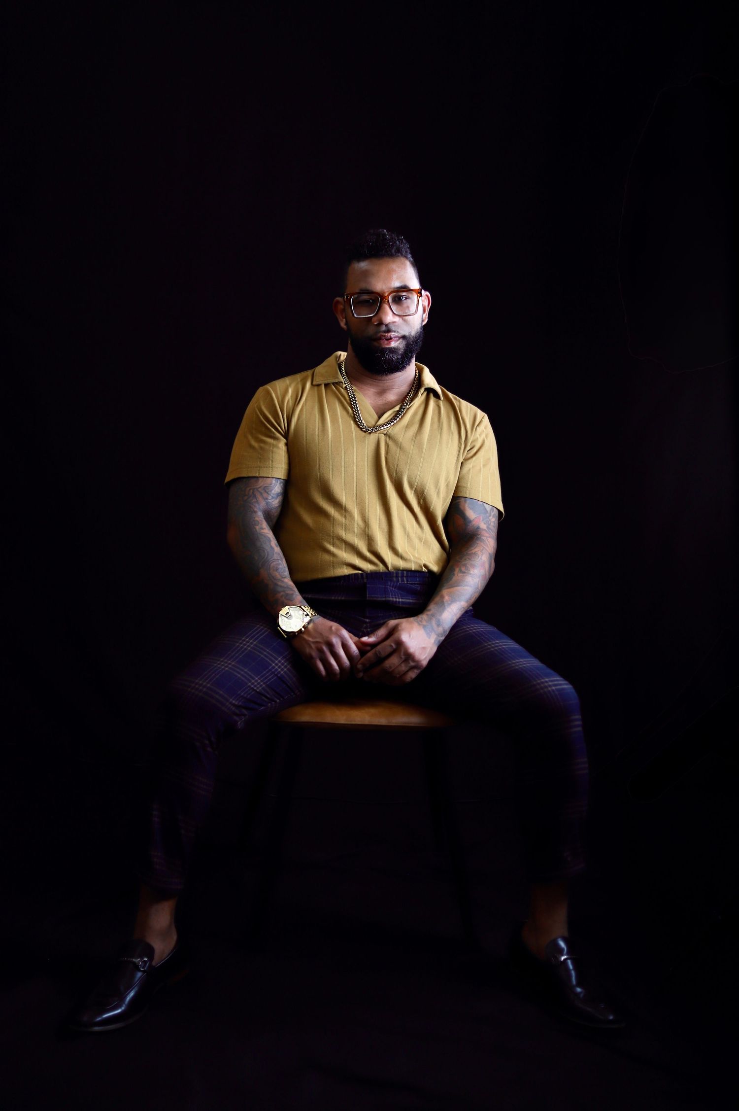 Man in yellow polo shirt and plaid pants sits on stool against black background, wearing glasses and gold watch.