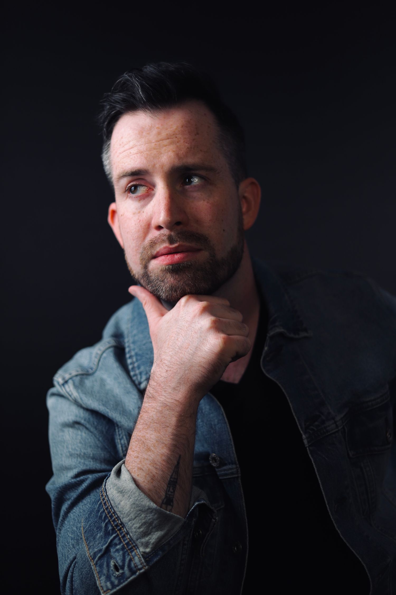 Professional portrait of a person in a denim jacket against a dark background with contemplative pose.
