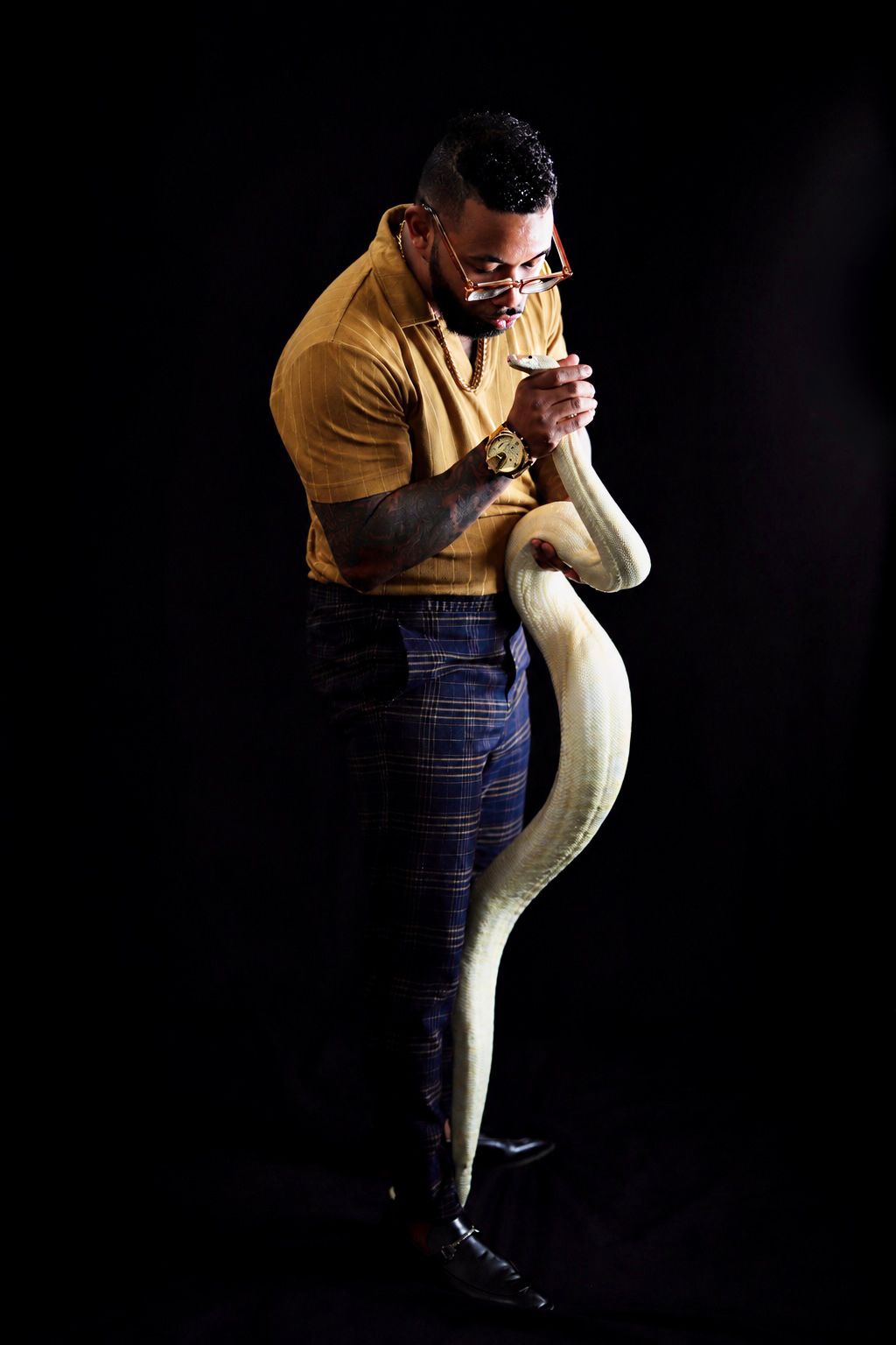 A muscular man in a yellow shirt and plaid pants holds a large albino python snake against a black background.