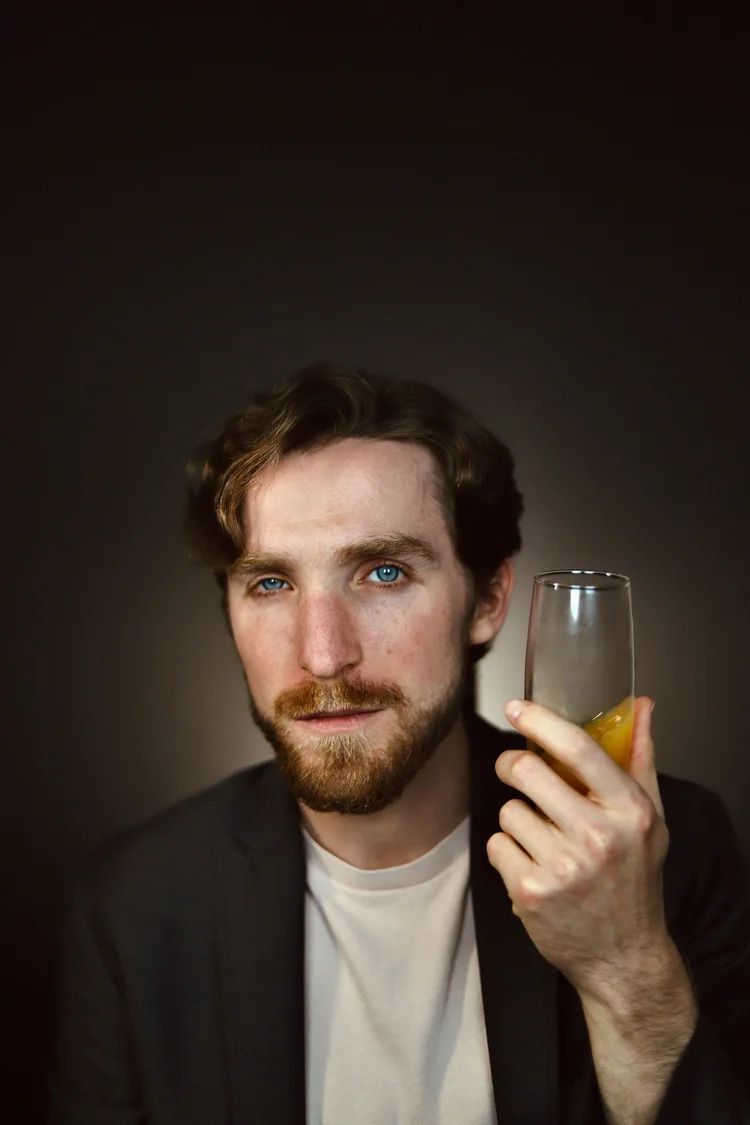 Moody portrait of a bearded person holding a glass with orange garnish against dark background.