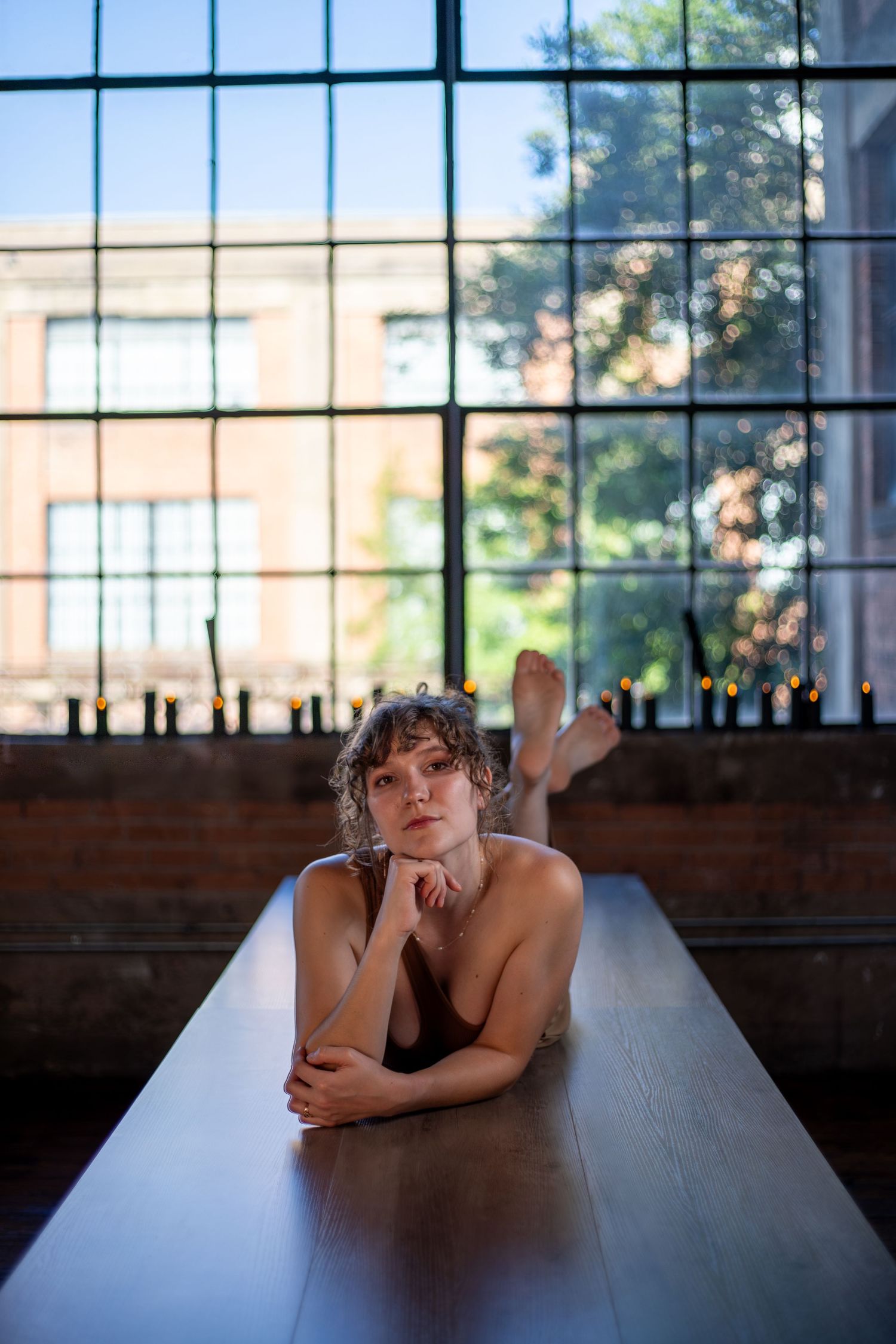 Young person with curly hair resting on a long table near large industrial windows with candles and brick building outside.