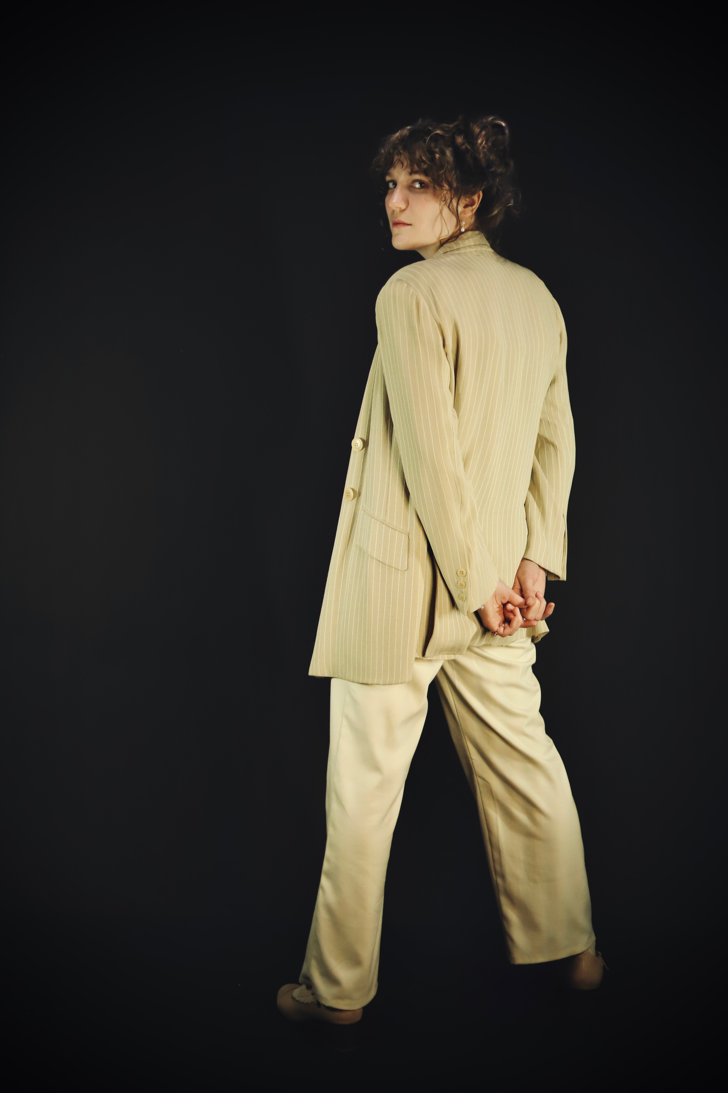 Figure in cream-colored oversized clothing poses dramatically against dark background in artistic photo series.