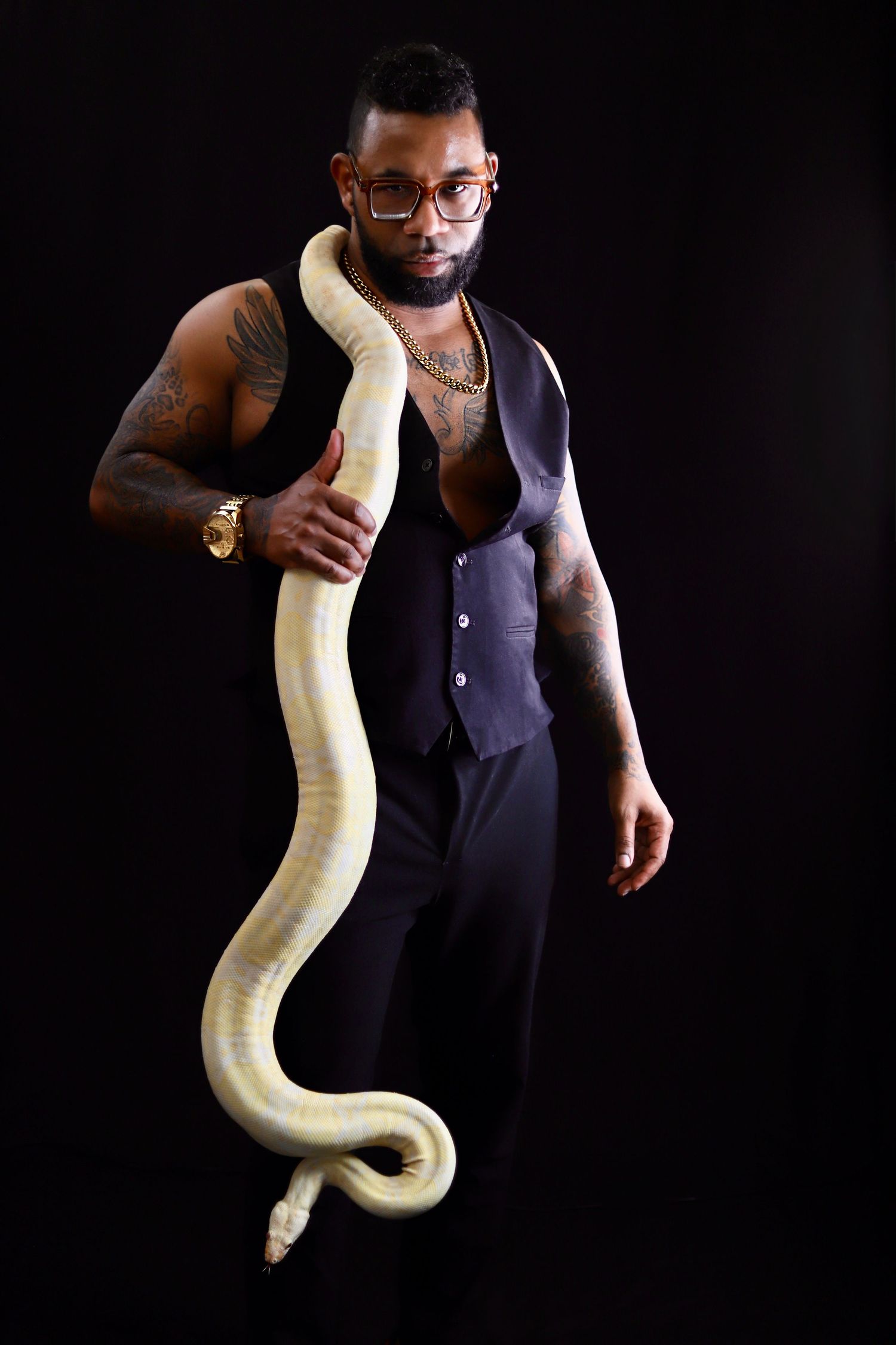 Tattooed man in black vest holding a large albino python snake, wearing glasses and gold jewelry against dark background.