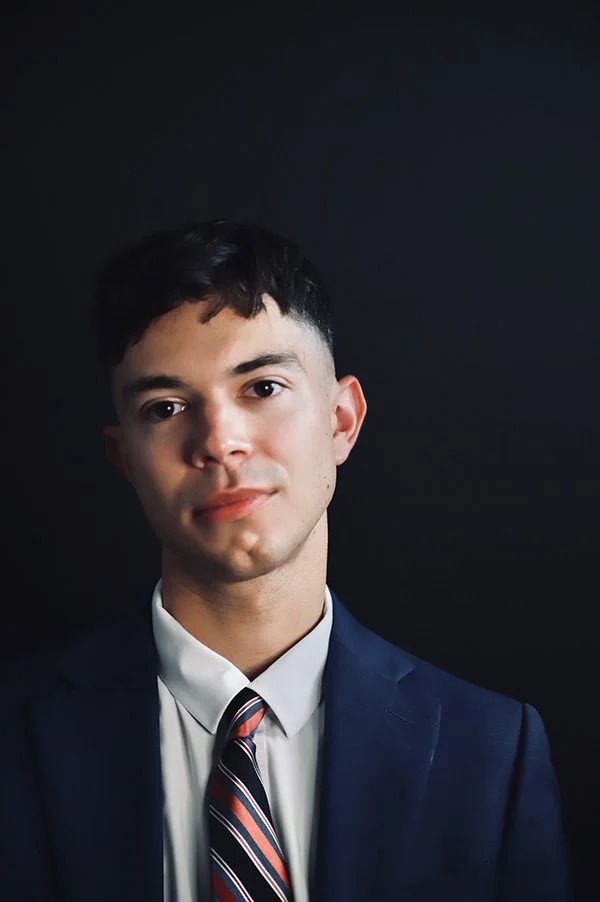 Professional headshot in navy suit and striped tie against dark background.
