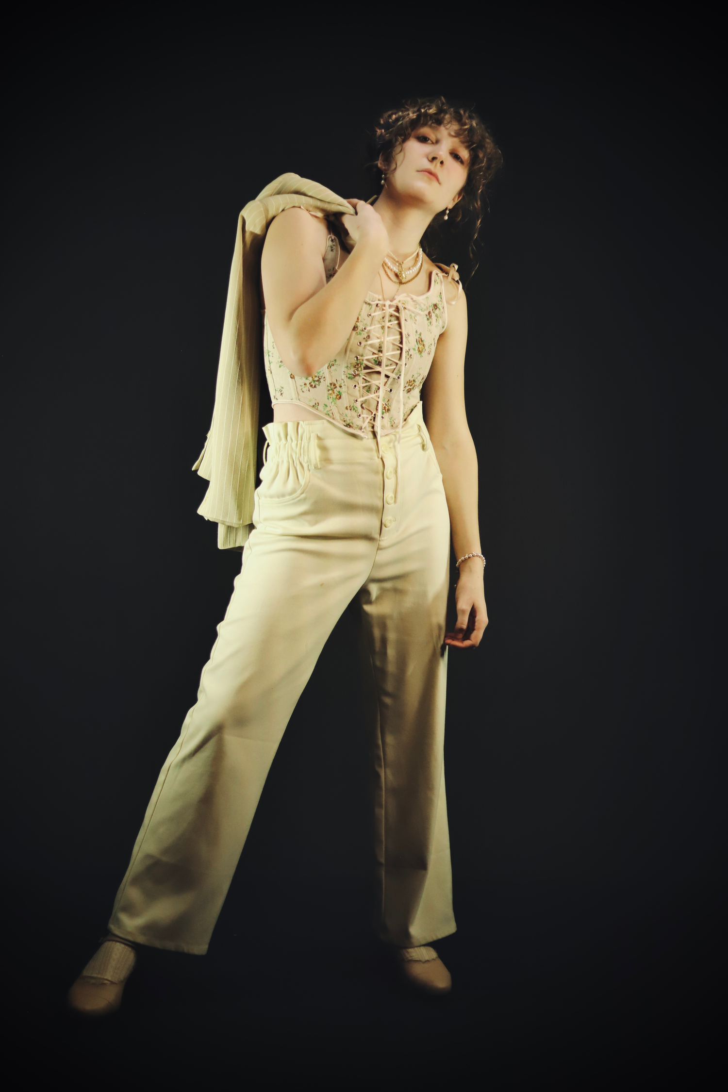Figure in cream-colored outfit with high-waisted pants and lace top poses dramatically against dark background.