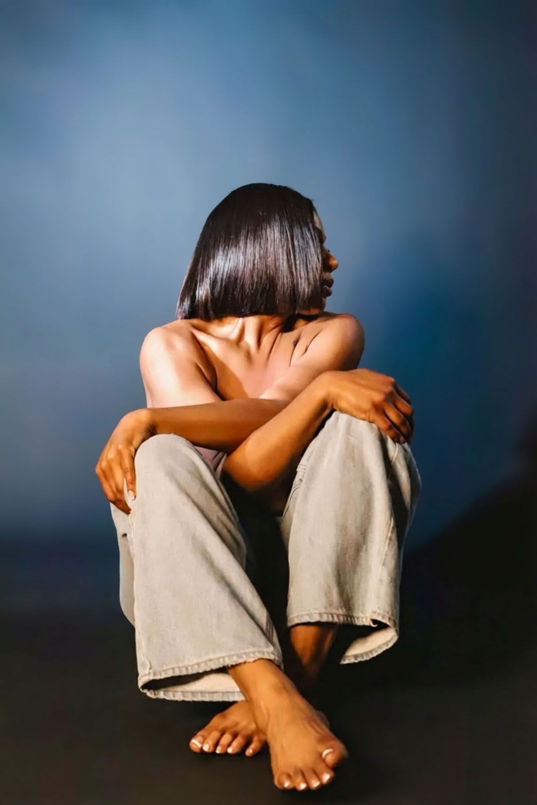 A person in distress sits curled up with head down and knees pulled to chest against dark background, showing vulnerability.