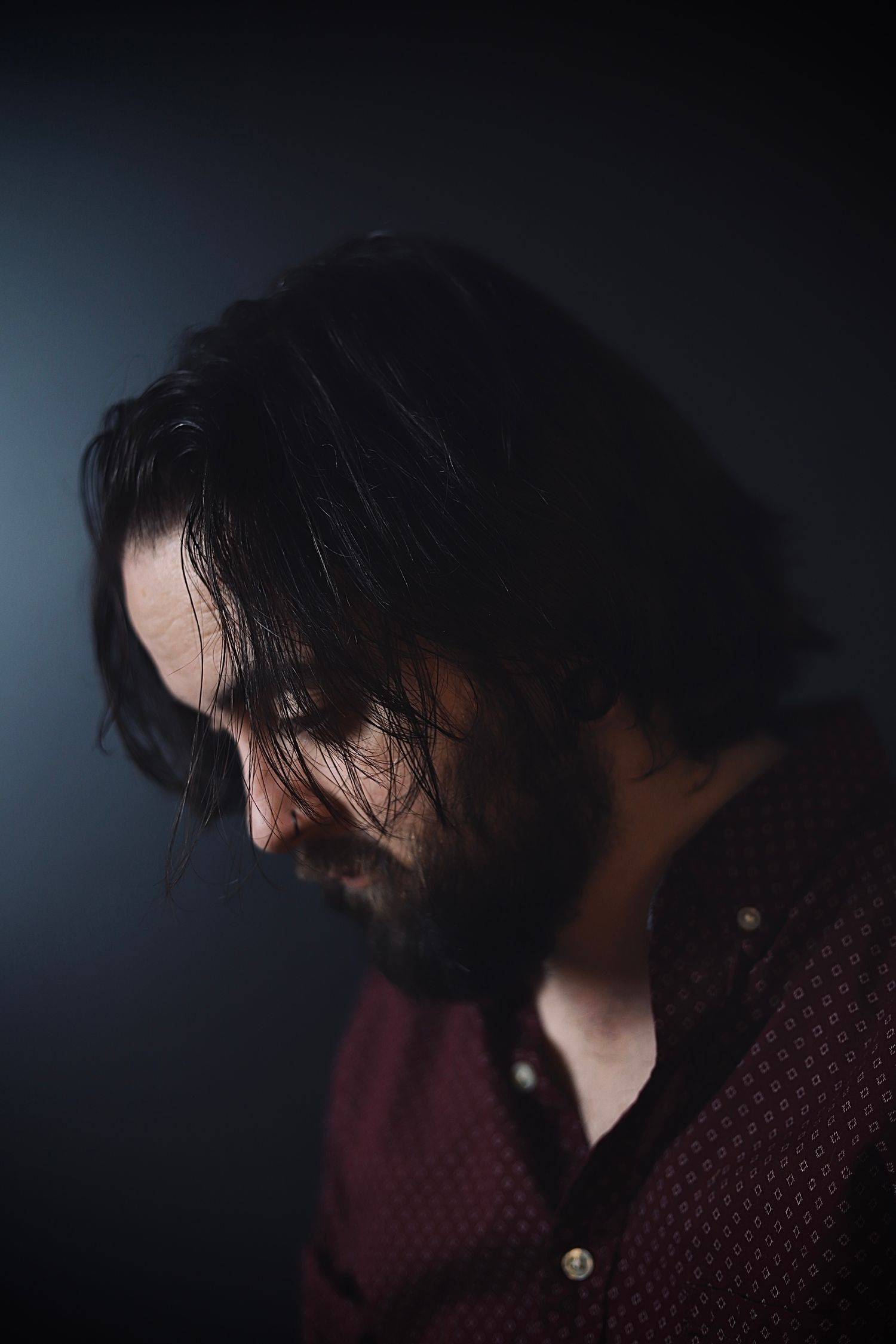 Dramatic low-key portrait of a bearded person in a burgundy sweater looking down against dark background.
