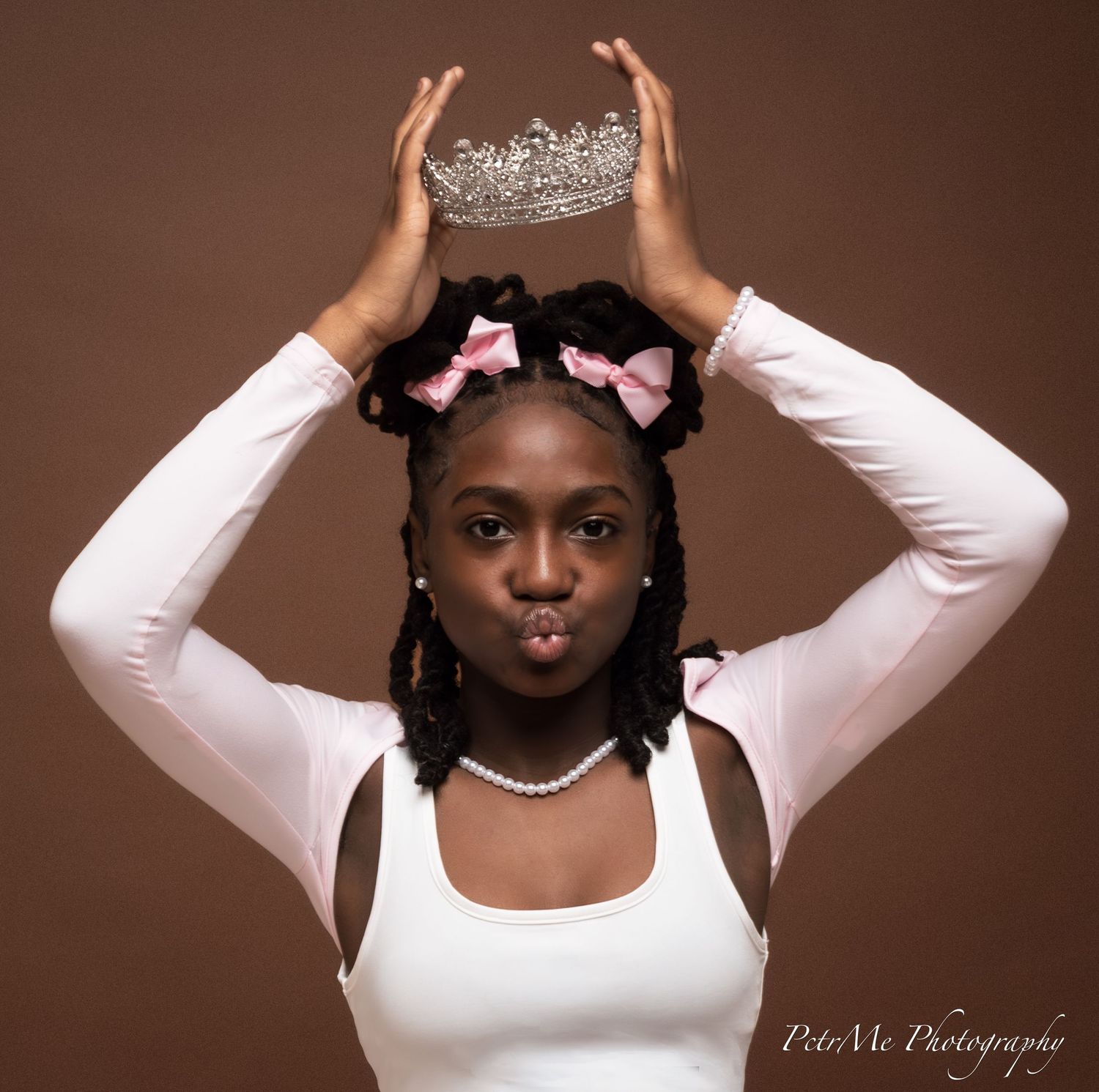 Studio portrait with a crown held overhead, wearing a white top, pink hair bows and pearl necklace against brown backdrop.