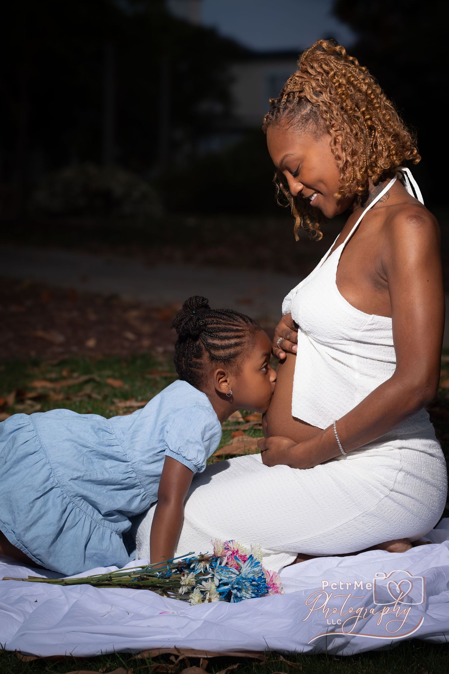 Pregnant woman in white dress sits outdoors while young girl kisses her baby bump during a maternity photo session.