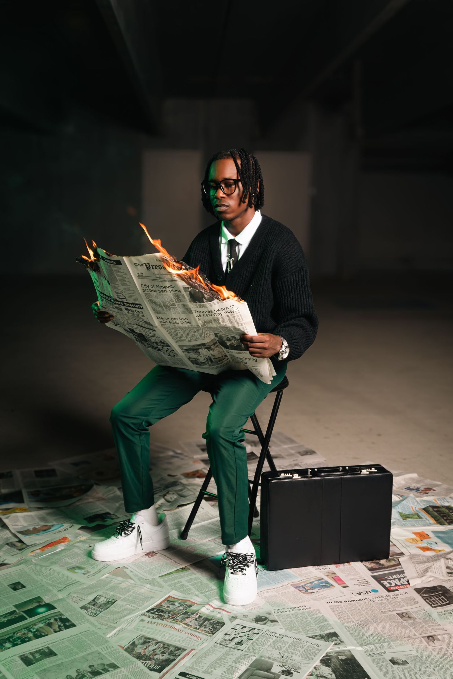 A stylish man in green pants reads a burning newspaper while seated, with a briefcase beside him on scattered papers.