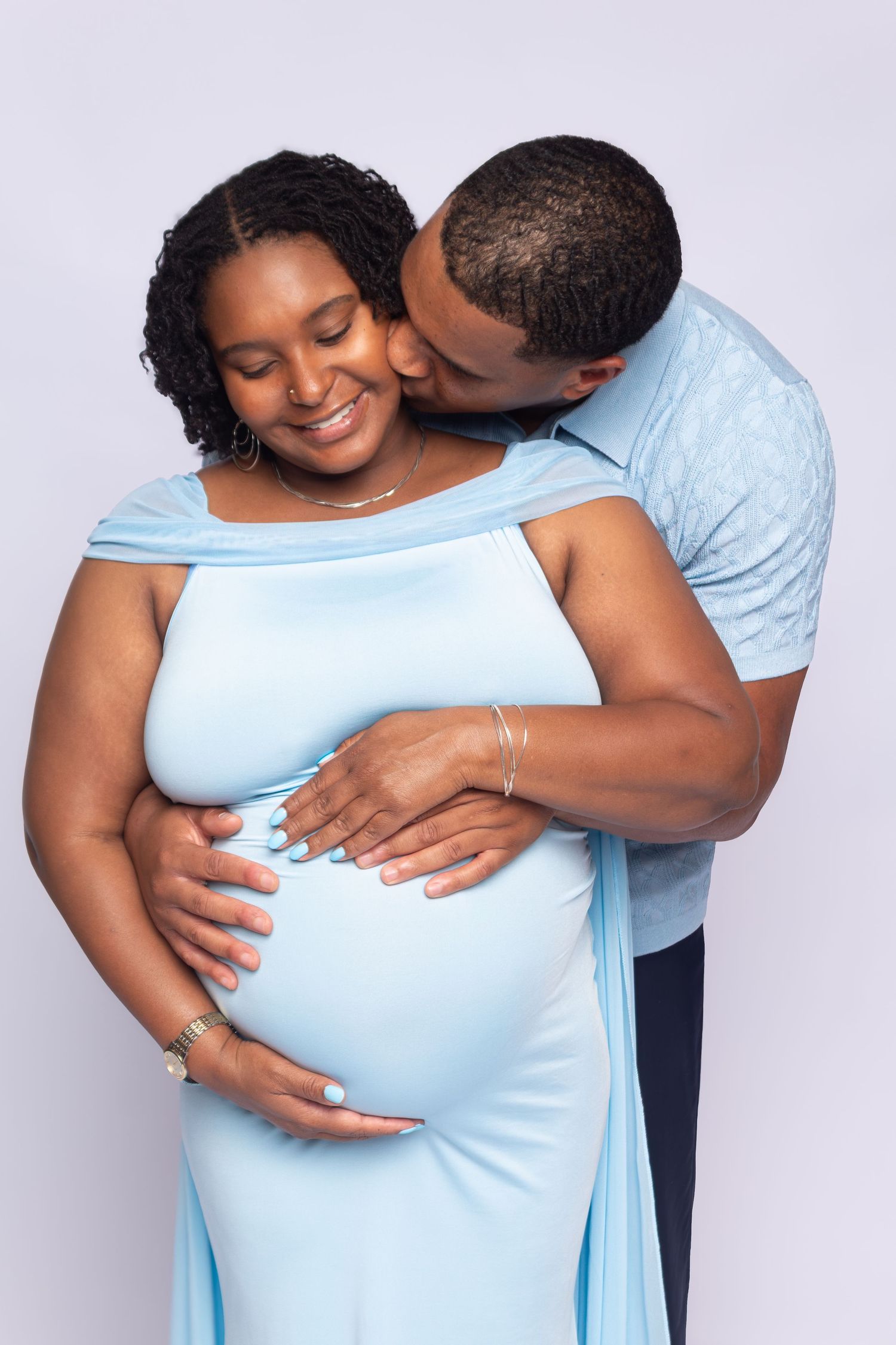 A man tenderly kisses a smiling pregnant woman in a light blue gown while embracing her baby bump.