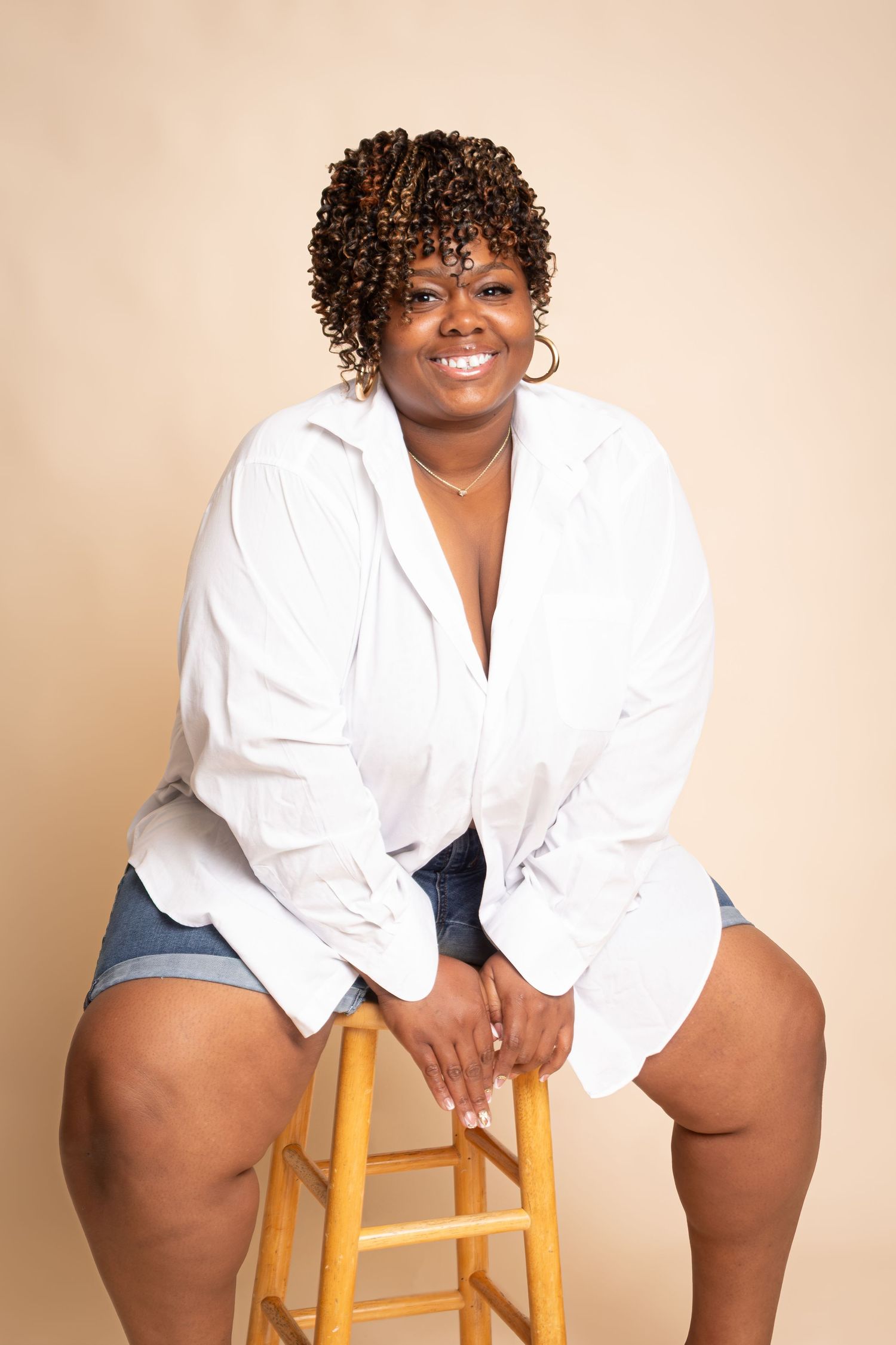 Smiling plus-size woman in white shirt and denim shorts sitting on a wooden stool against a beige background.