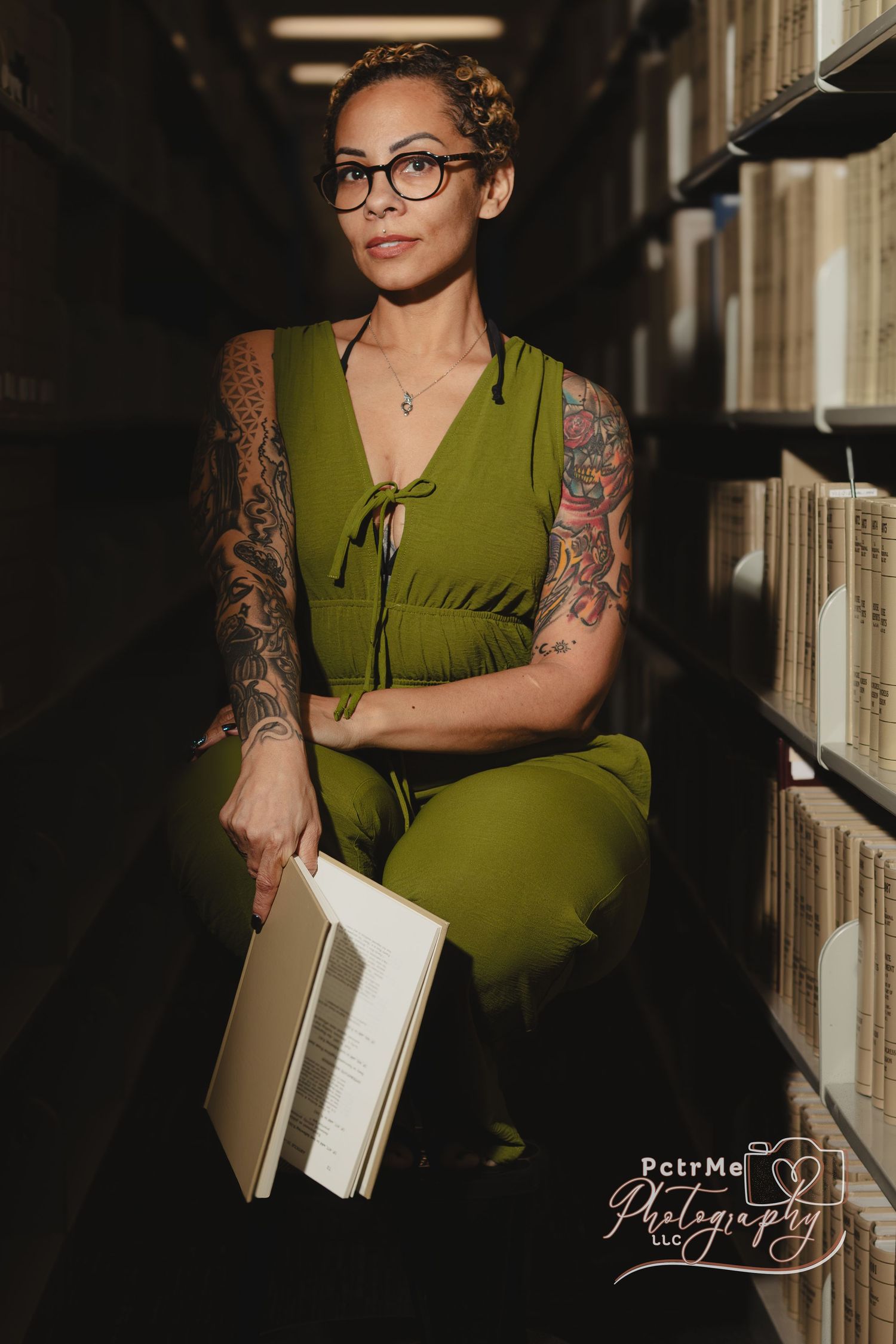 Tattooed woman in green jumpsuit holding a book between library shelves, wearing glasses in a moody portrait.