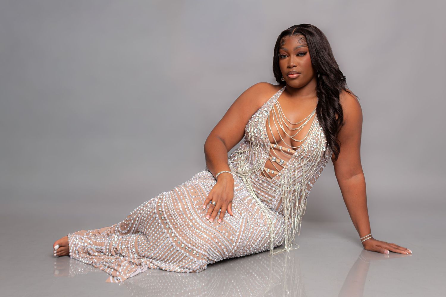 A plus-size woman poses elegantly in a beaded fringe gown on a gray studio backdrop.
