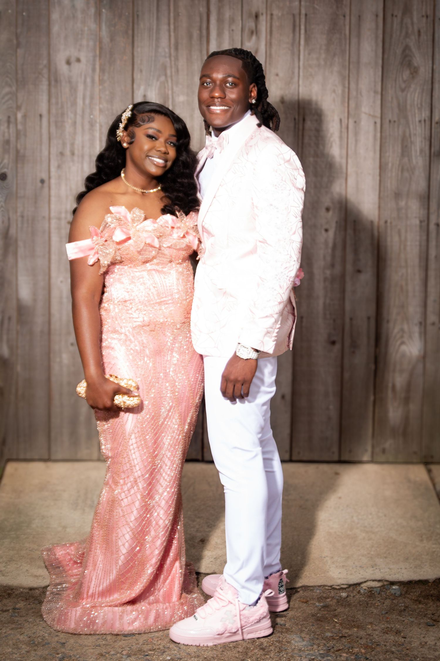 A couple dressed in matching pink formal wear poses together for a prom photo in front of a rustic wooden backdrop.