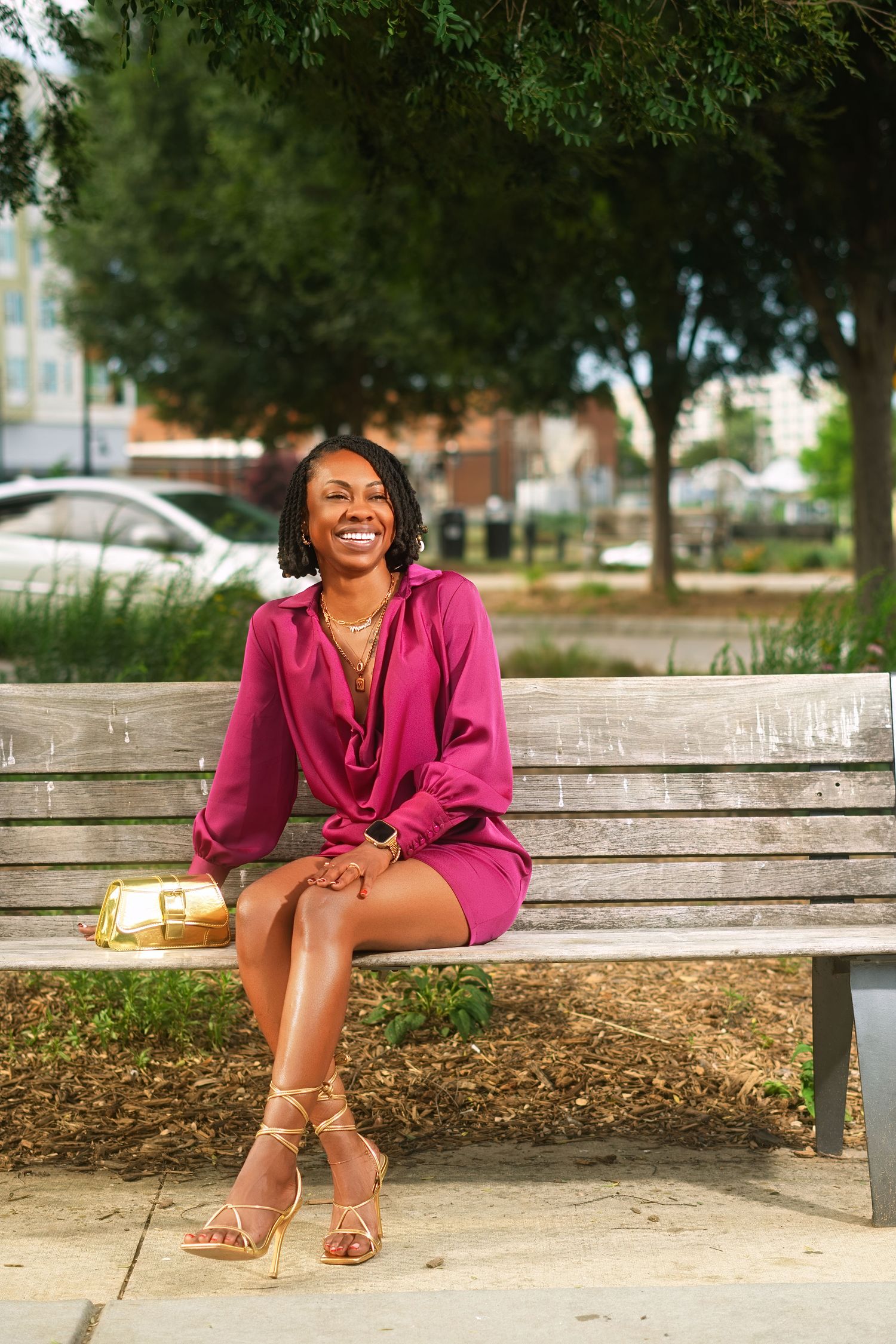 Smiling woman in magenta dress and gold heels sitting on a park bench with a gold clutch purse.