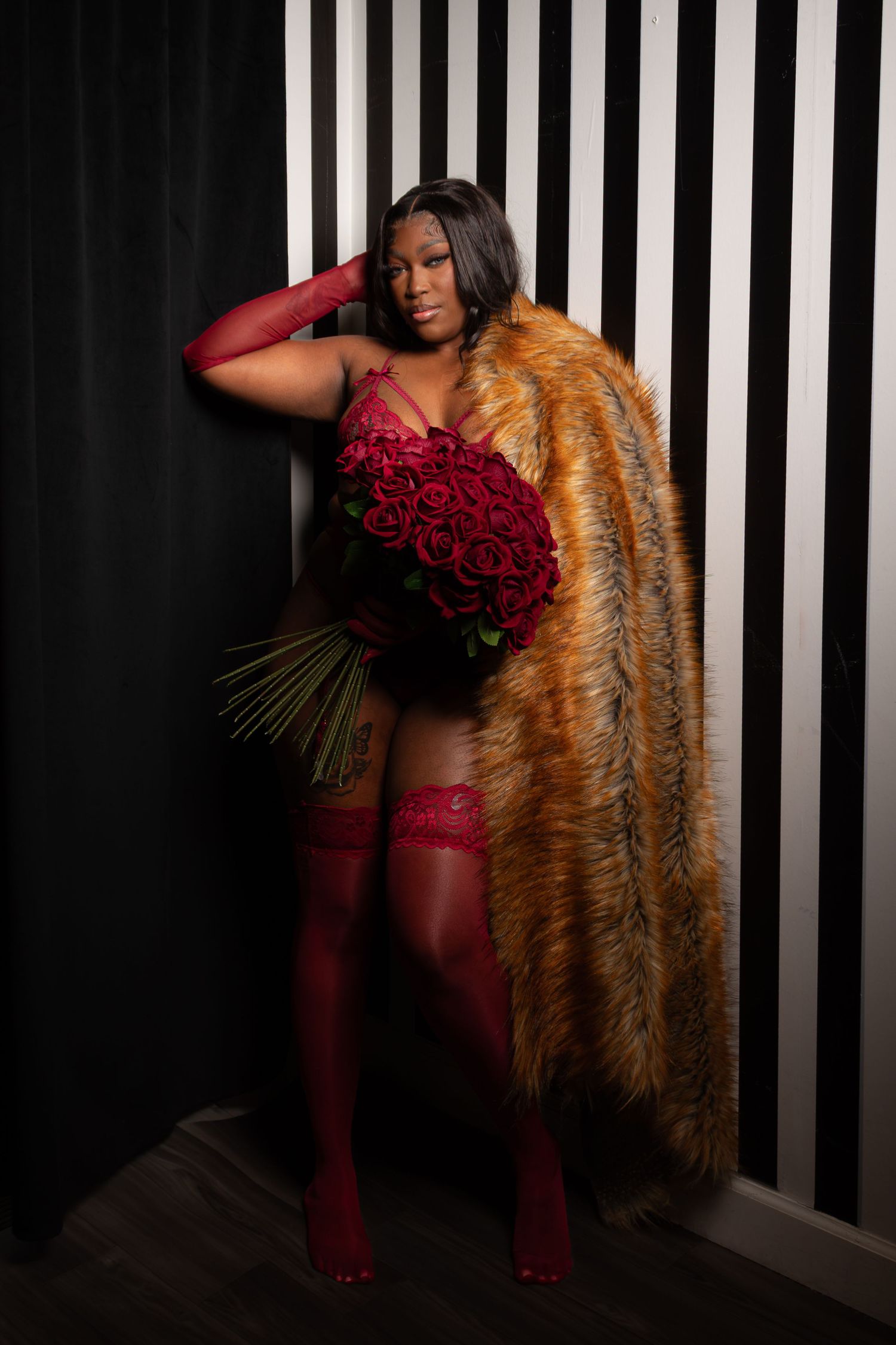A woman in red lingerie and fur coat holds red roses against a black and white striped backdrop.