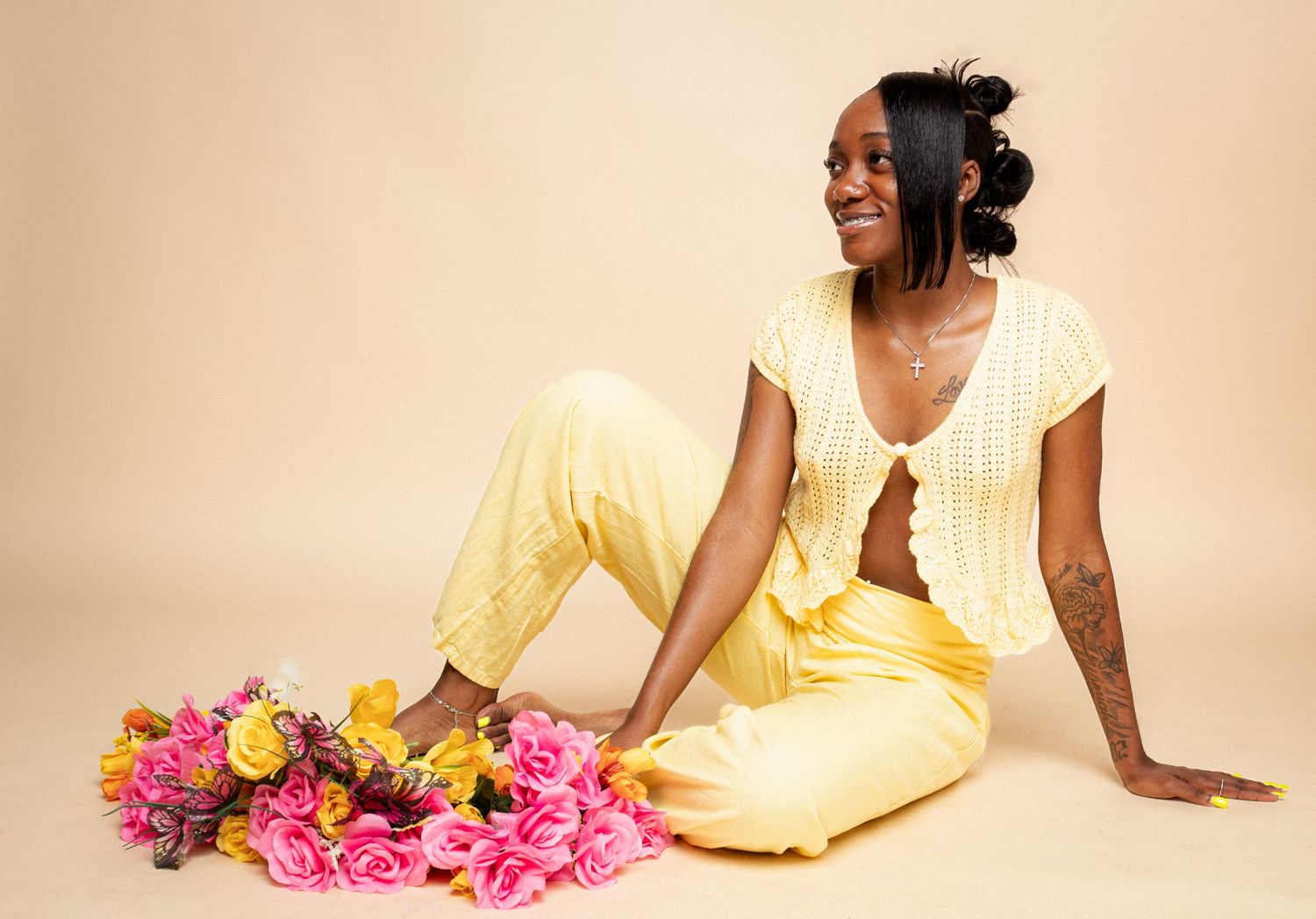 Woman in yellow outfit sitting with pink and yellow flowers, smiling, on beige background.