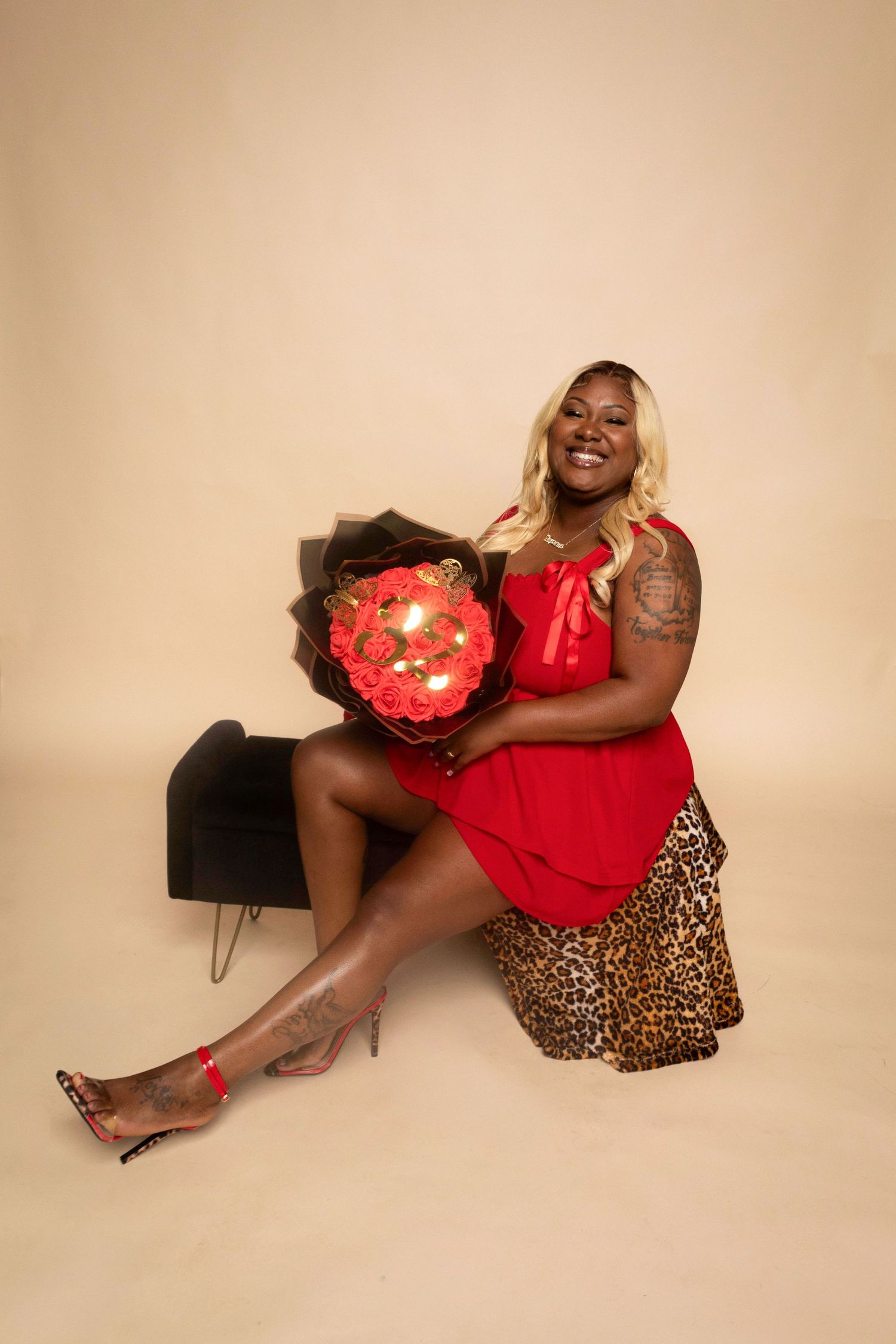 A person wearing red and leopard print poses with a heart-shaped pillow in a studio setting against a beige backdrop.