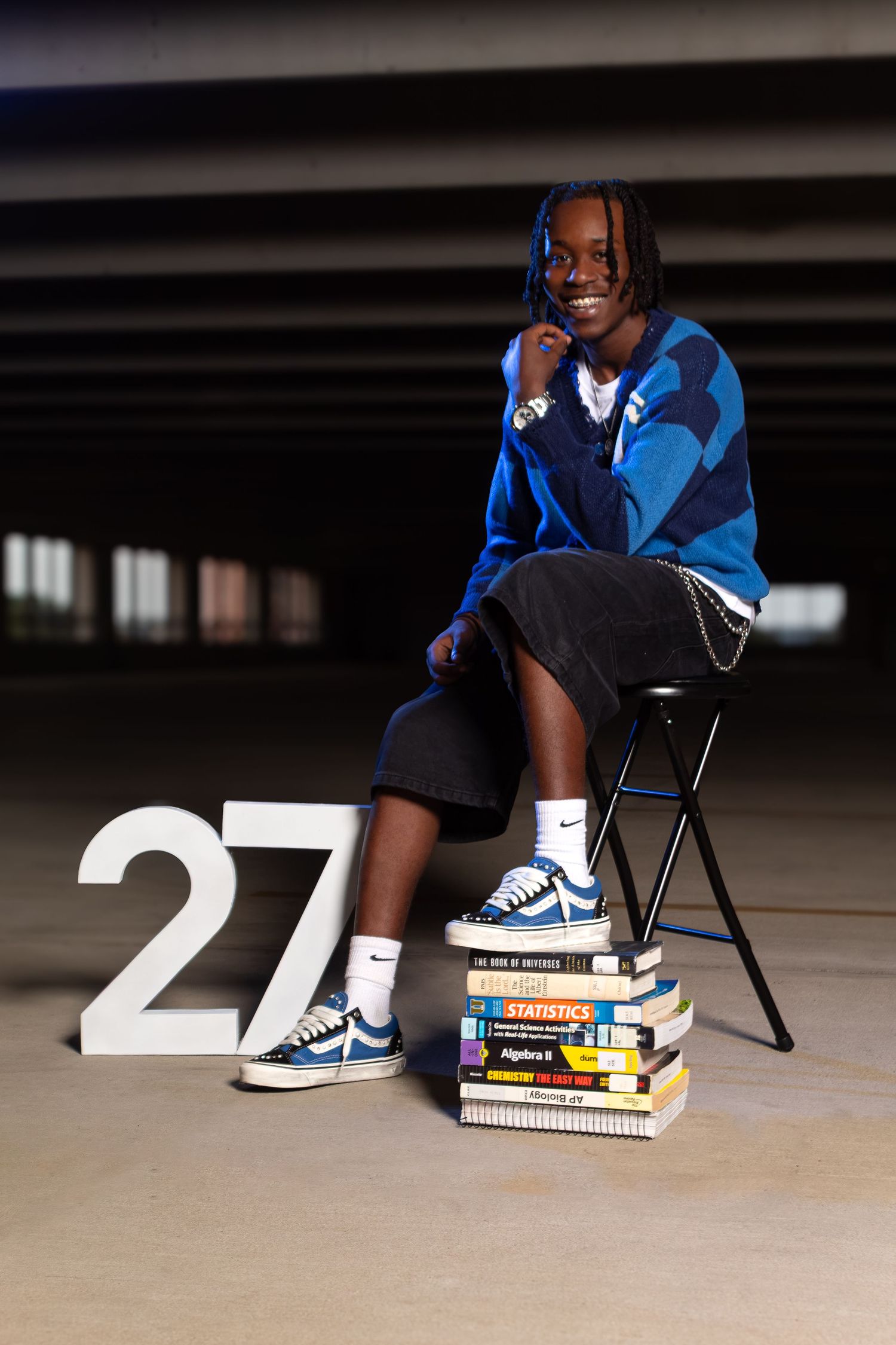 Student sits on stool beside number 27, resting feet on stacked textbooks, wearing blue Vans and blue hoodie.