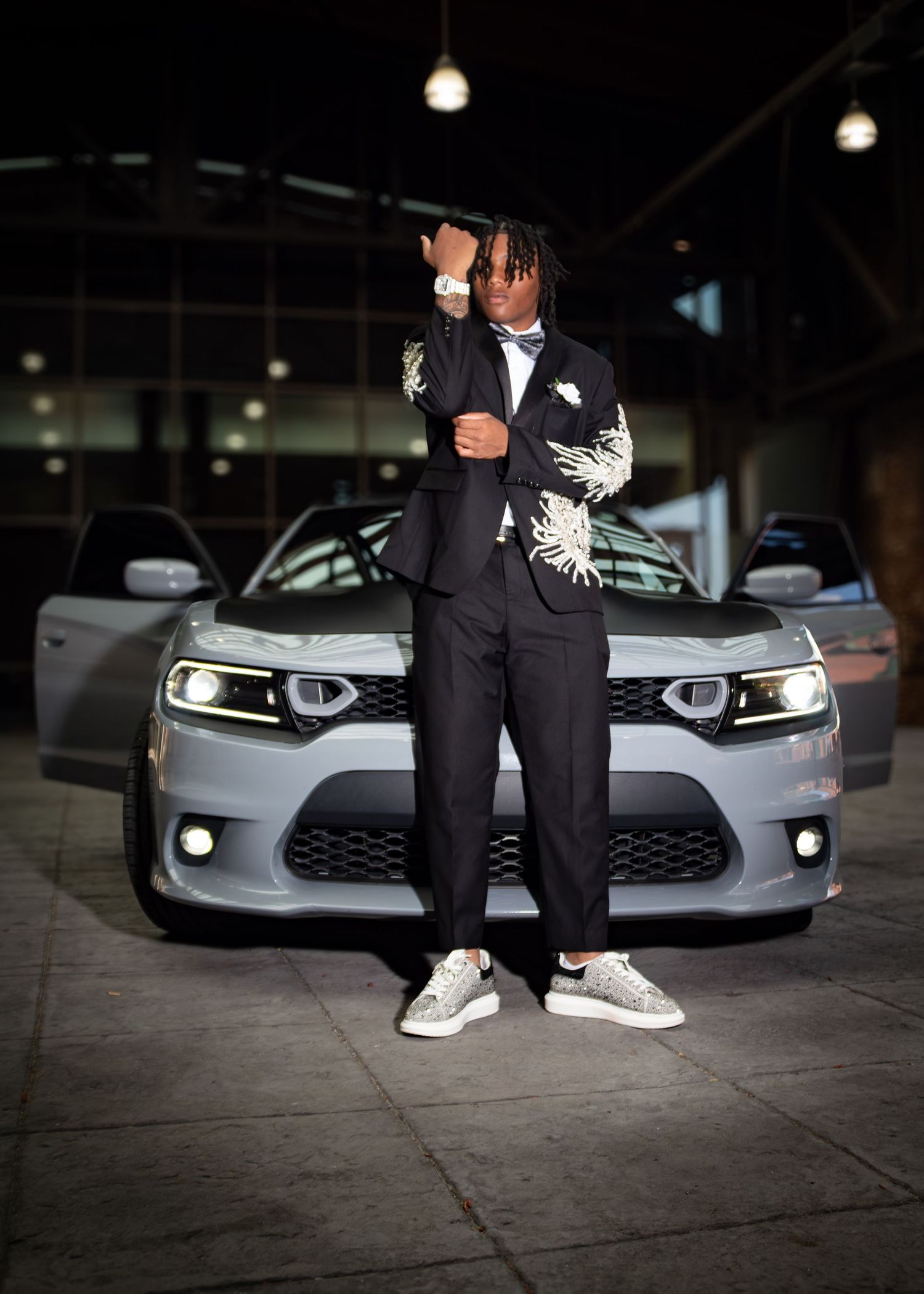 Young man in floral blazer poses confidently in front of silver Dodge Charger in dimly lit showroom.