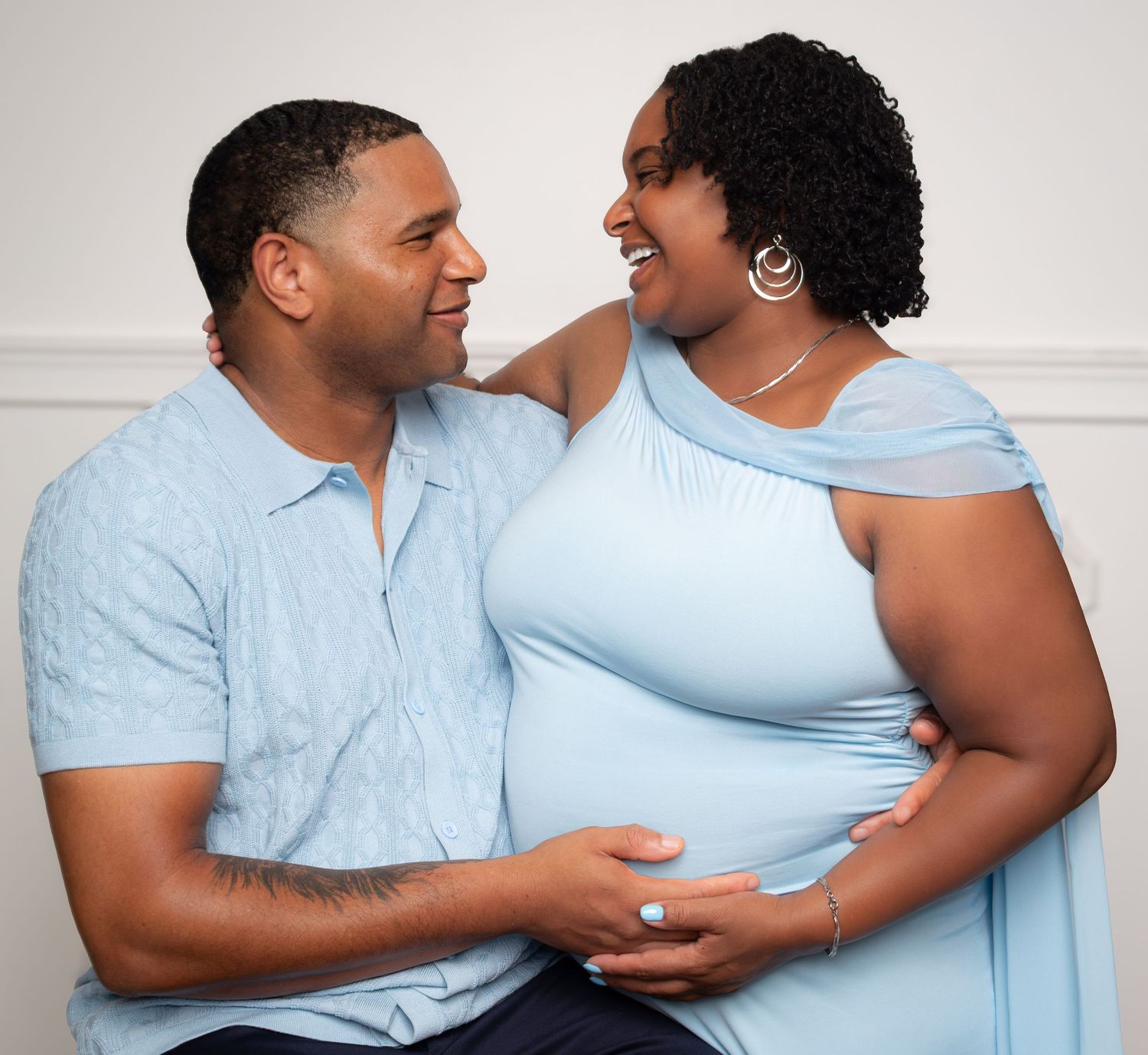 A couple in matching light blue outfits share a joyful moment during a maternity photoshoot, with the man gently holding her baby bump.