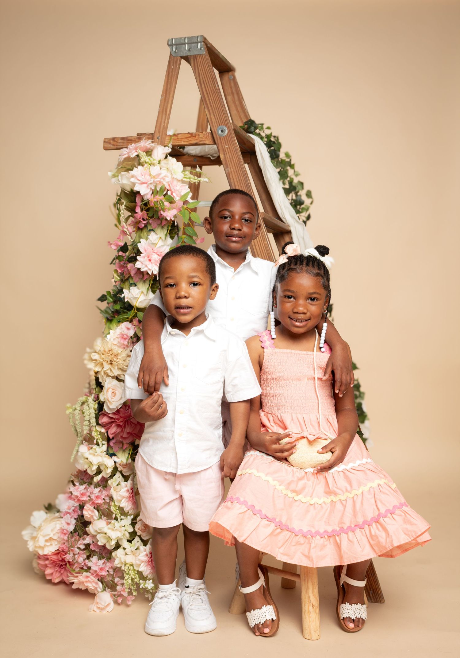 Three children pose in front of a floral-decorated wooden ladder, dressed in pastel spring outfits for a studio portrait.