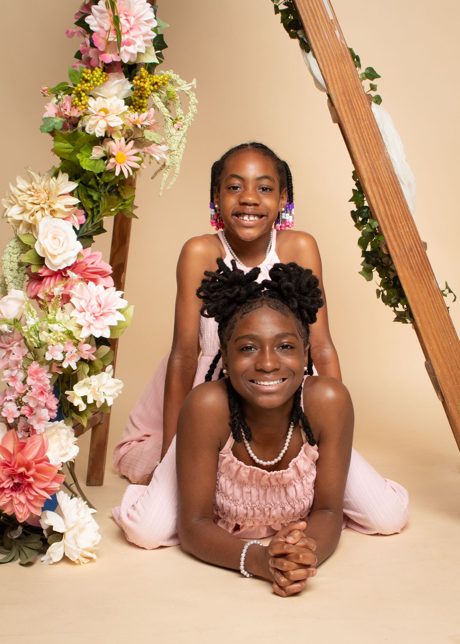 Two young girls pose together in a floral studio setting, wearing pink outfits with pearl accessories.