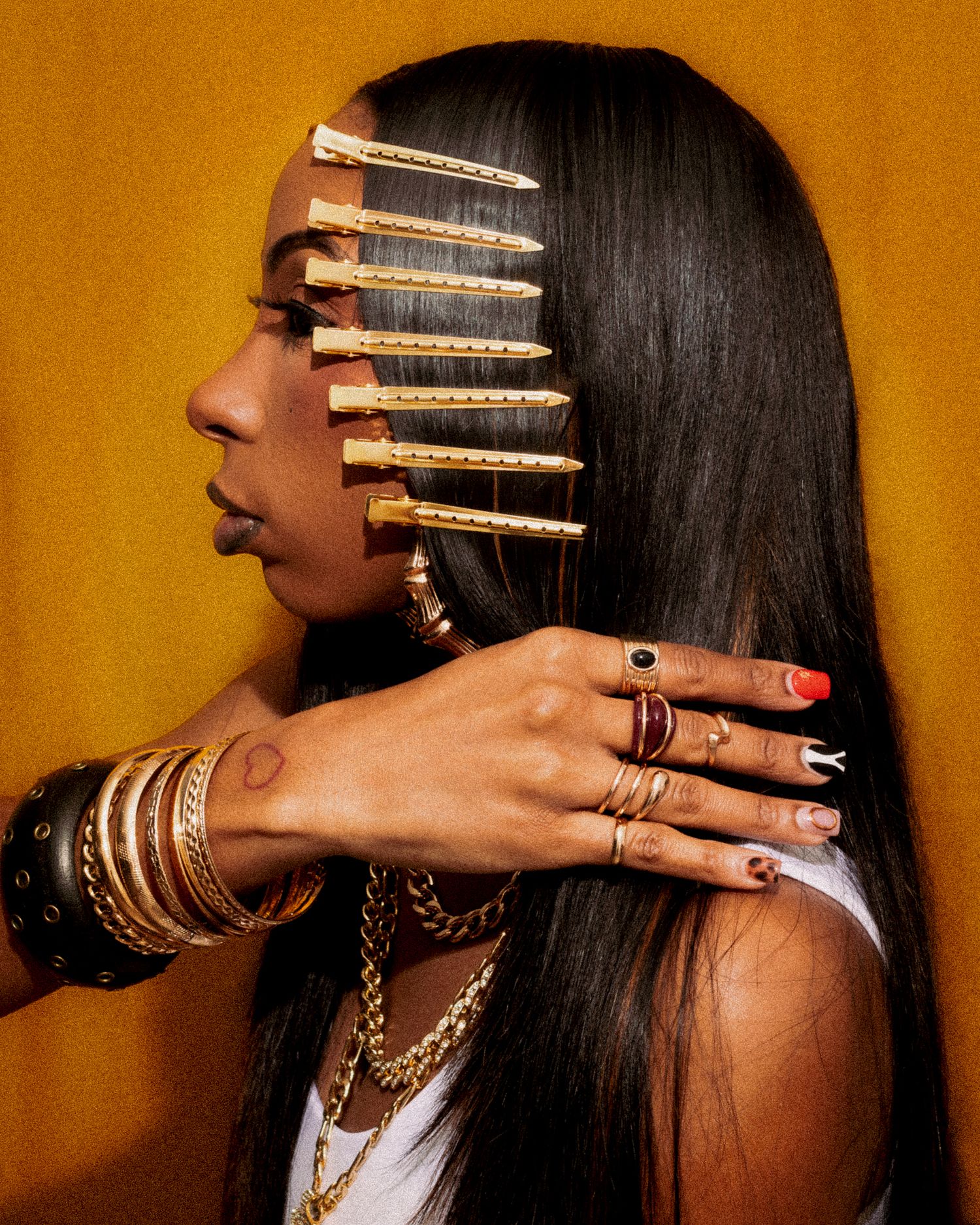 Woman with gold hair clips, layered gold necklaces, bangles, and colorful rings poses against yellow background.