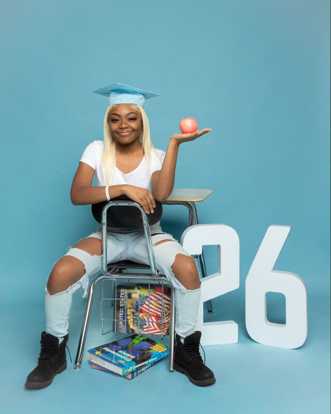 Graduate in cap and white outfit sits on chair with apple and books against blue wall with '26' numbers.